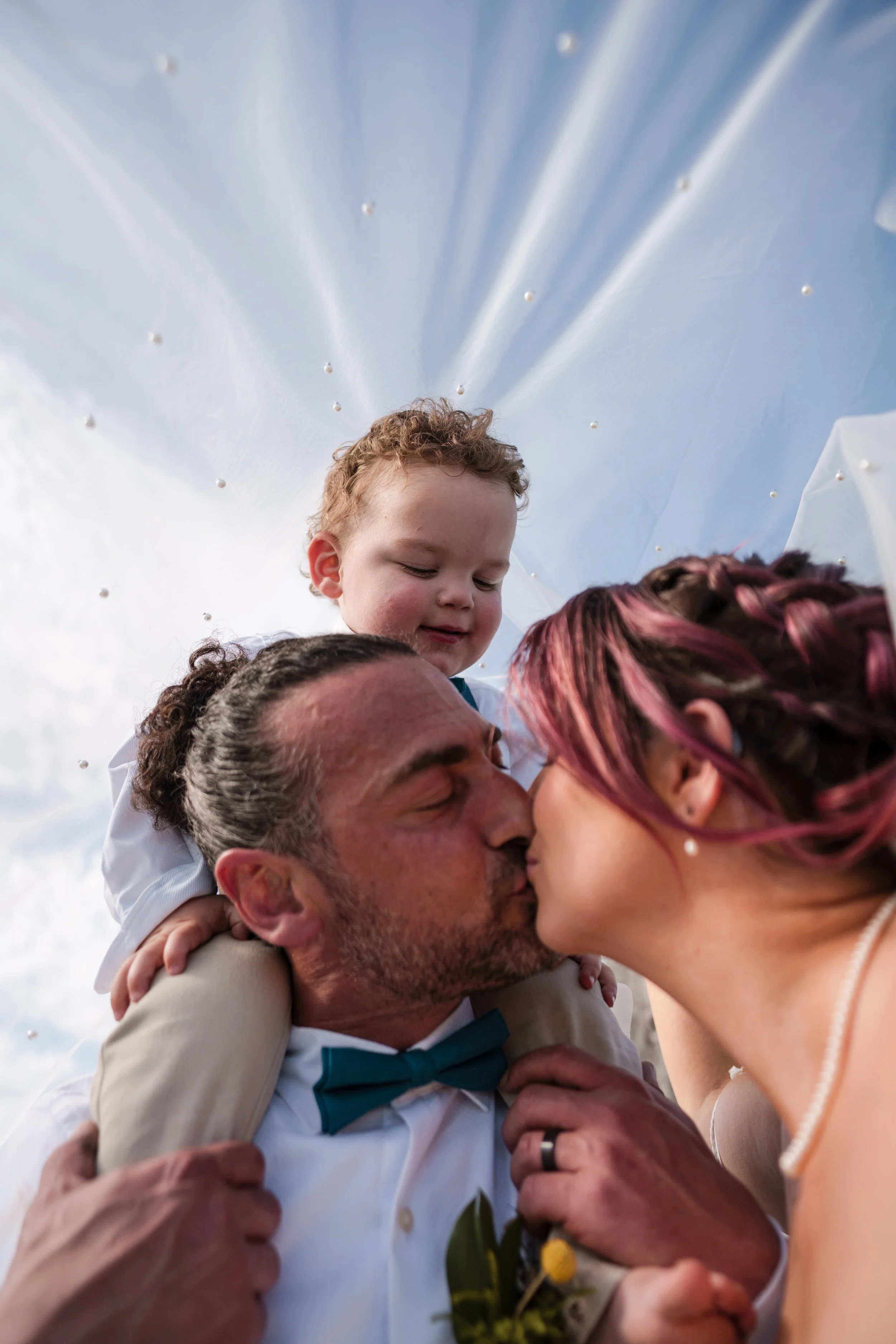 A family kiss at a wedding celebration, with a young boy sitting on a man's shoulders and a woman kissing the man, all outdoors with a blue sky background.