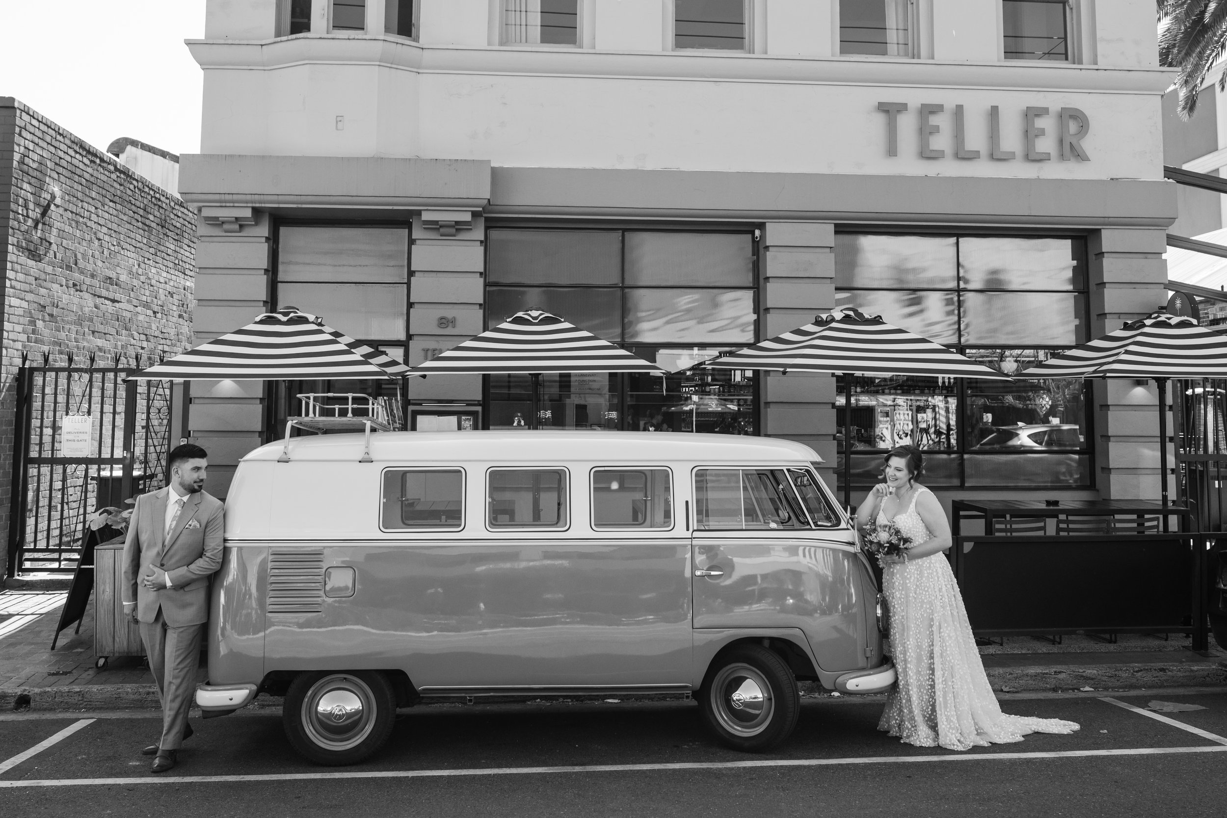 A black-and-white photo of a man in a suit leaning against a vintage van and a woman in a wedding dress holding a bouquet, standing by the van in front of a building with the sign 'TELLER'. The building has striped umbrellas outside.