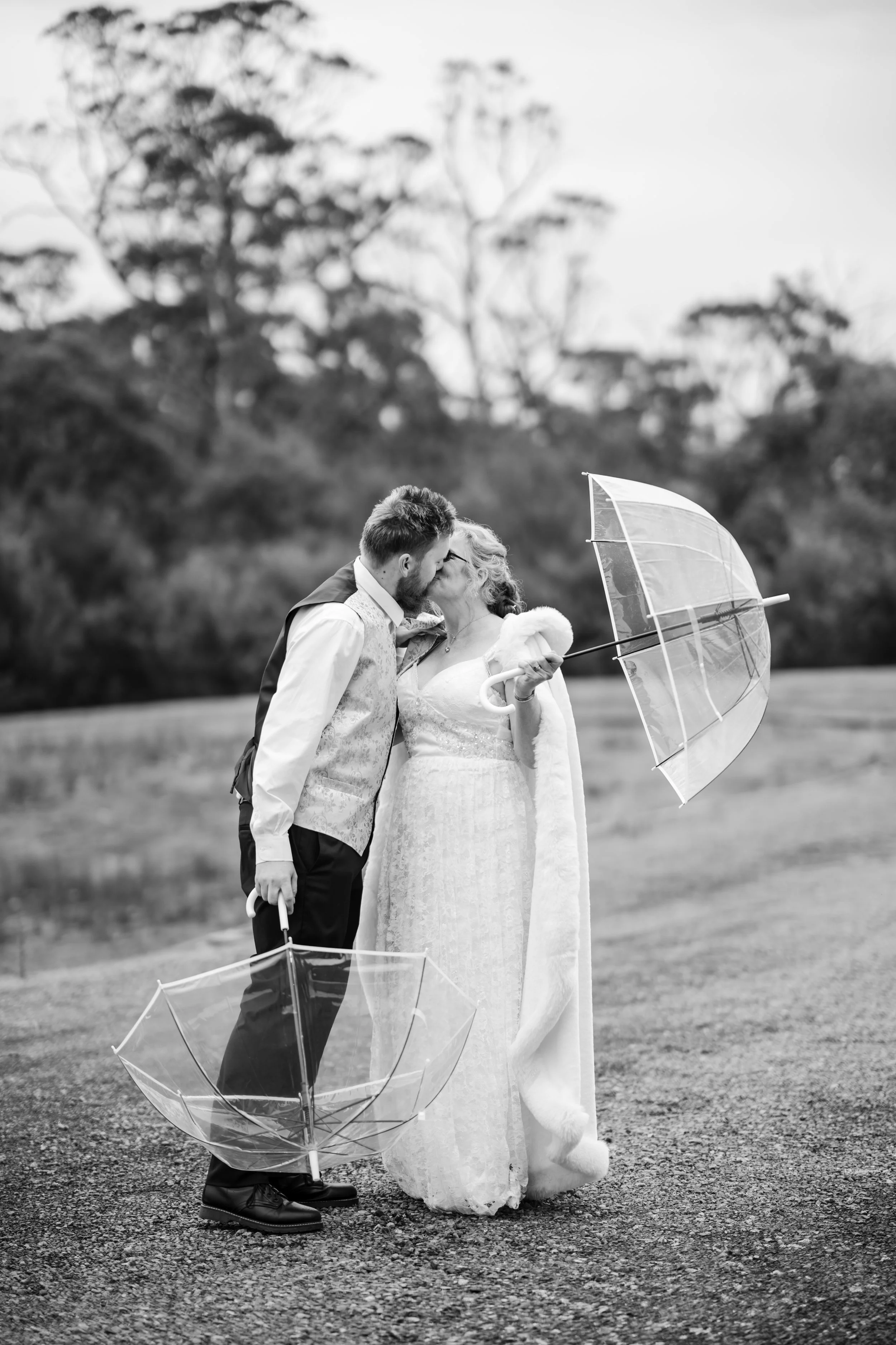 A couple in wedding attire sharing a kiss outdoors, holding umbrellas, with trees in the background.
