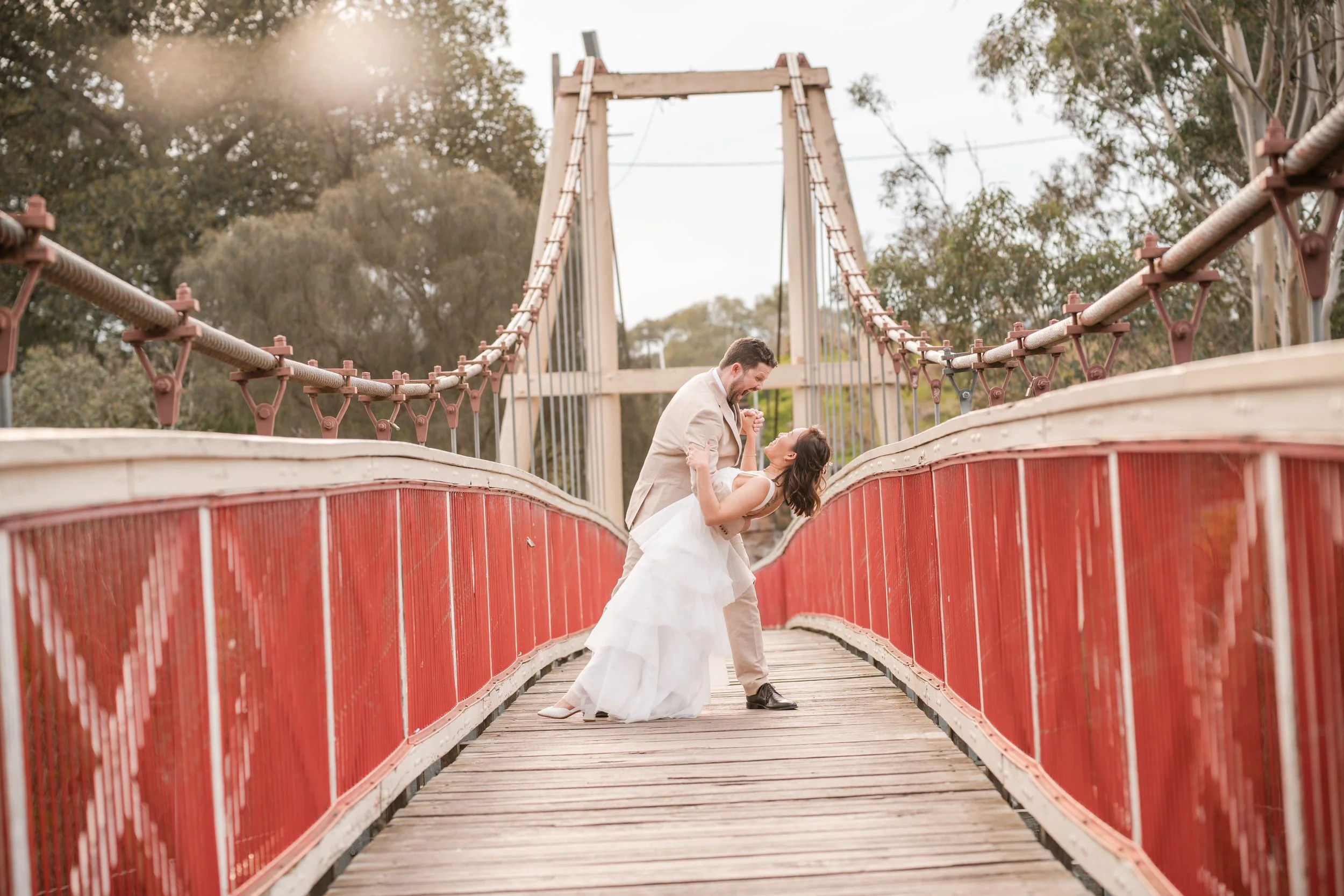 A couple in wedding attire dancing on a red bridge surrounded by trees.