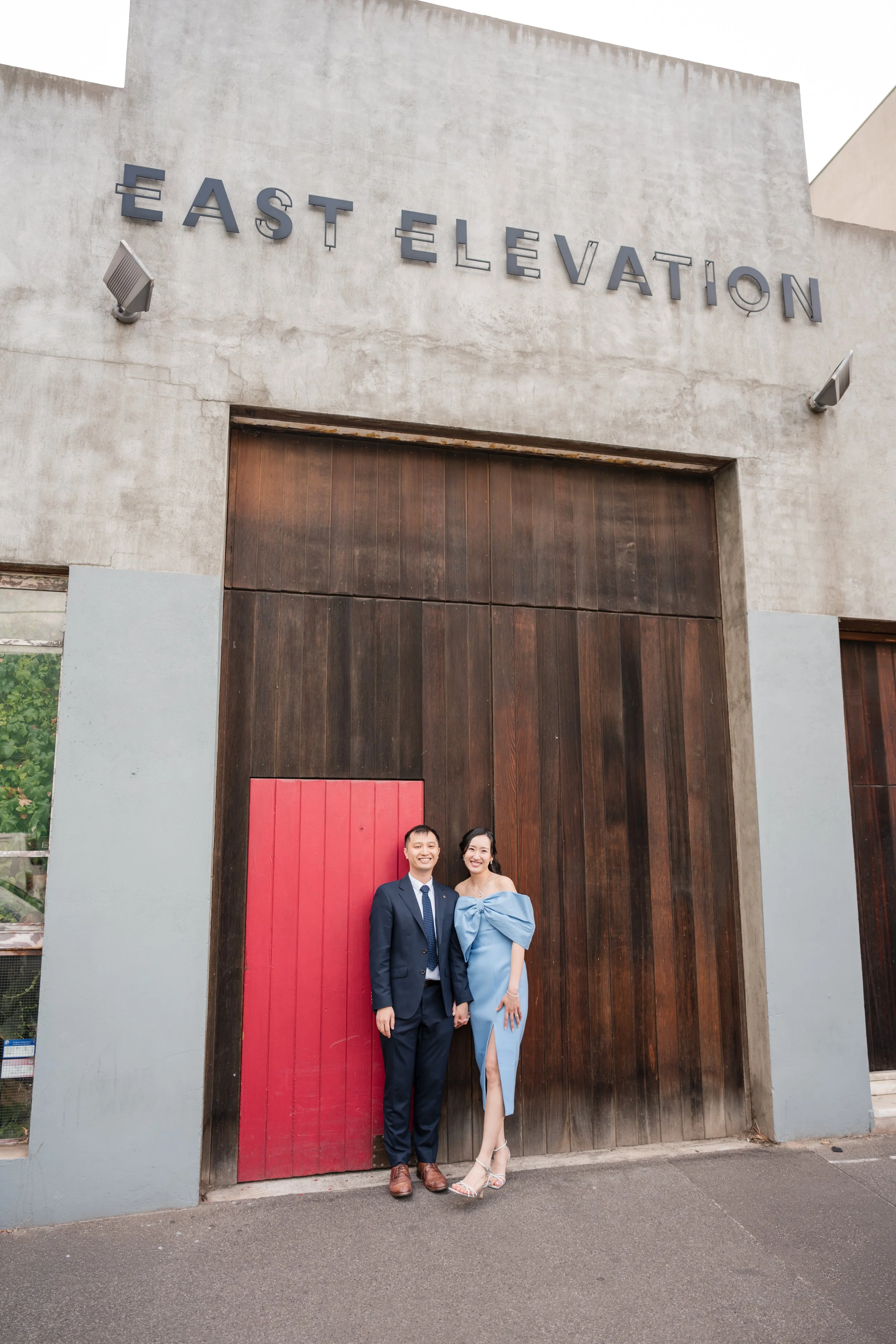 A smiling man and woman in formal attire standing in front of a large wooden door under a sign that reads 'EAST ELEVATION' on a concrete building.