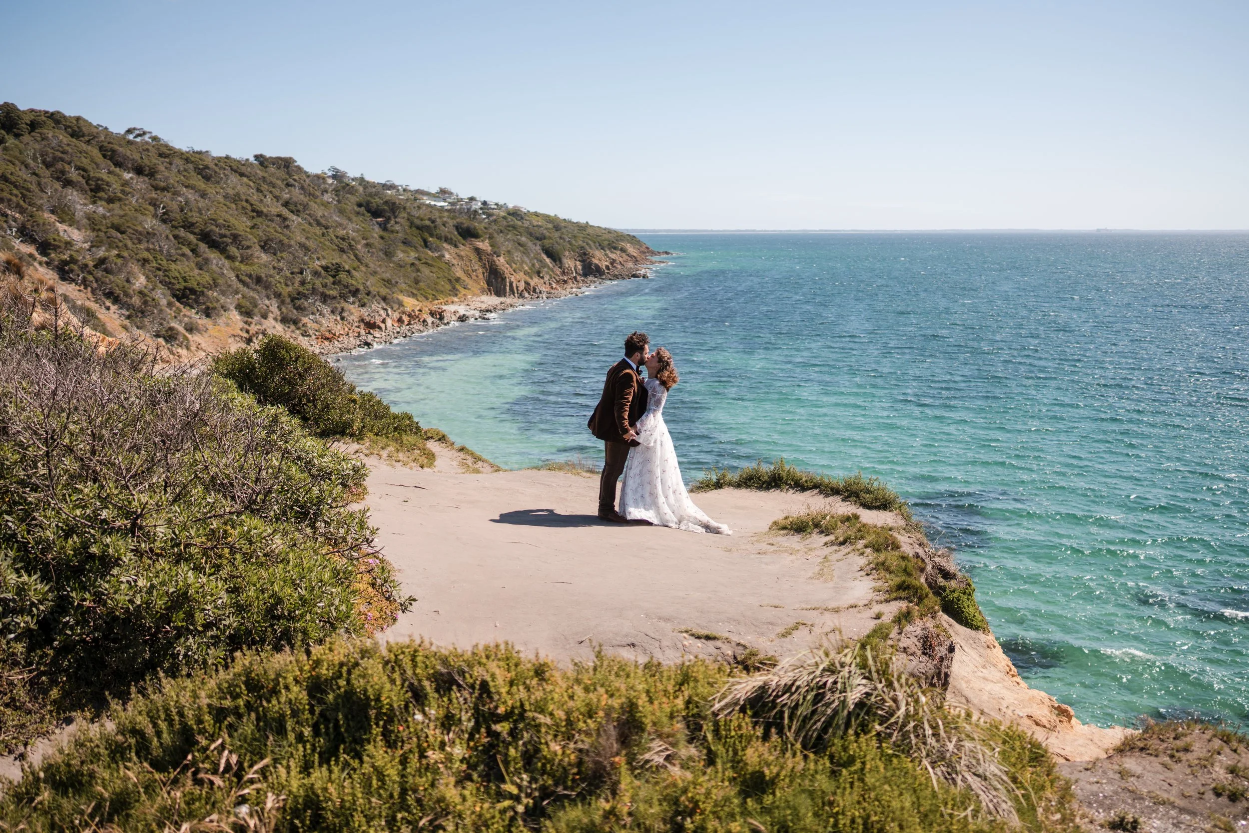 A couple dressed in wedding clothes, standing on a sandy cliff by the ocean, sharing a kiss with a scenic view of the sea and rolling hills in the background.