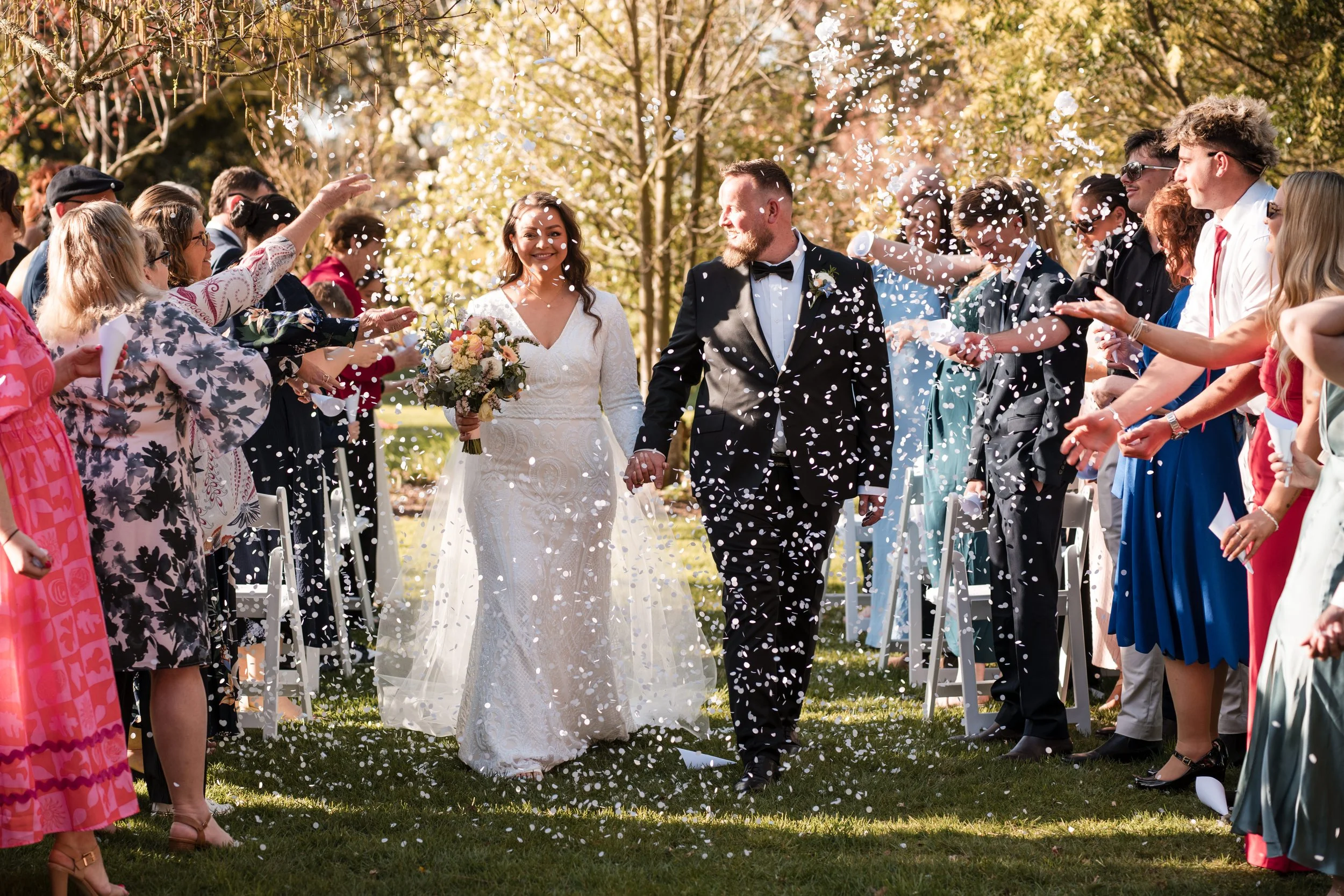 A bride and groom walking hand in hand through a confetti shower at their wedding ceremony, surrounded by friends and family in outdoor setting with trees and sunshine.
