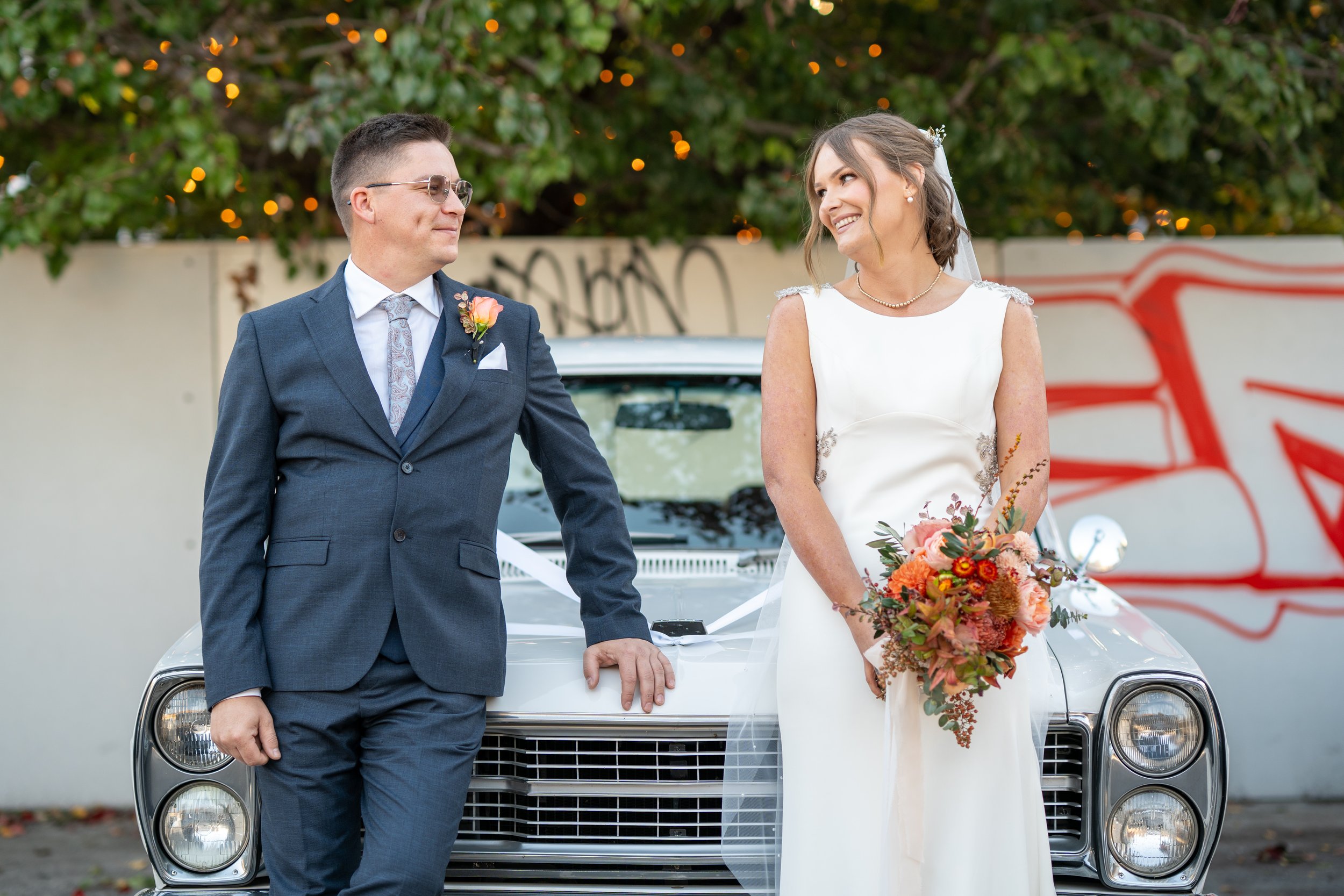 A bride and groom stand in front of a silver car with ribbons, smiling at each other outdoors, with a graffiti wall and trees with string lights in the background.