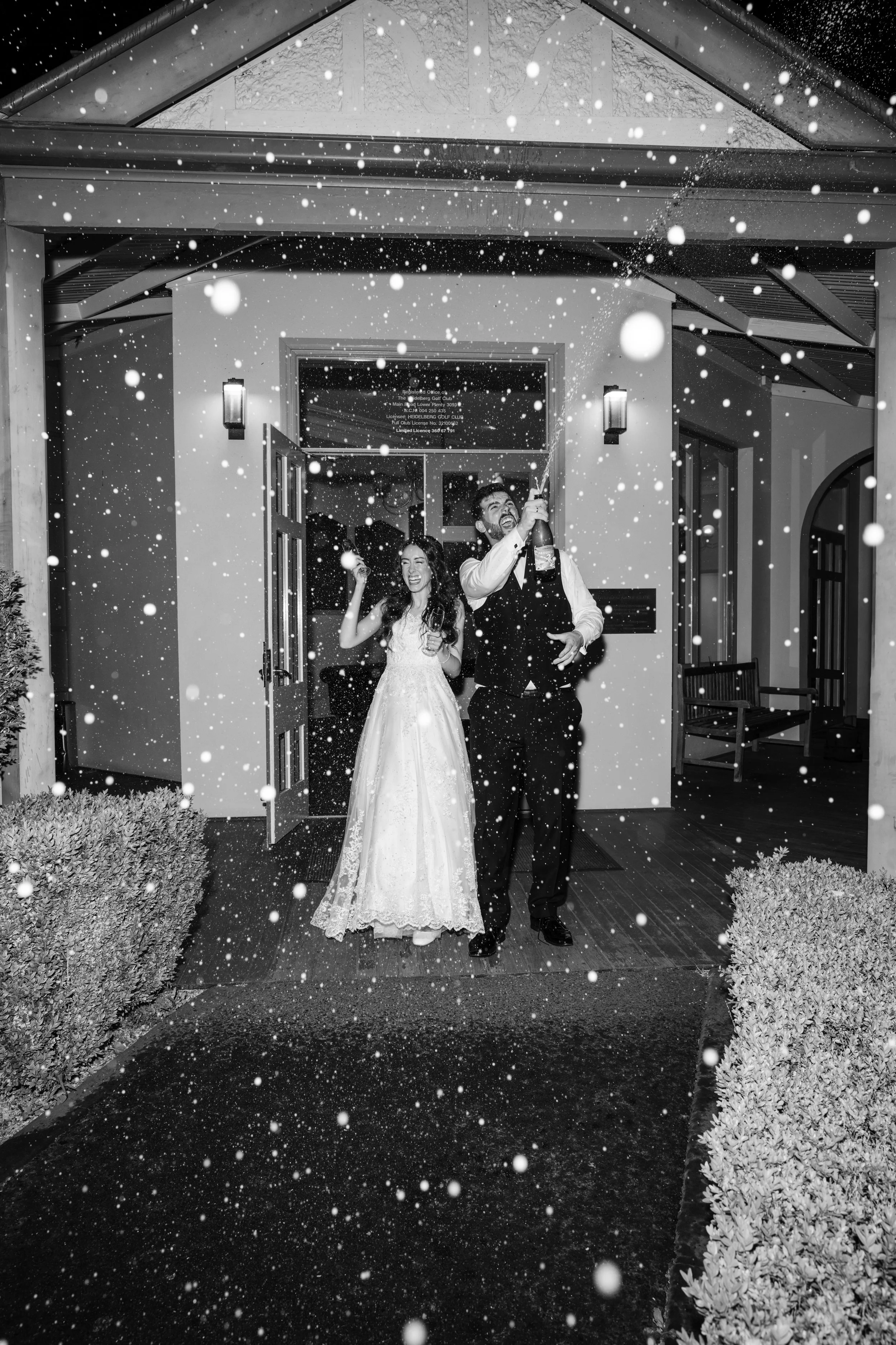 Black and white photo of a newlywed couple in wedding attire celebrating outside a building at night, with snow falling around them.