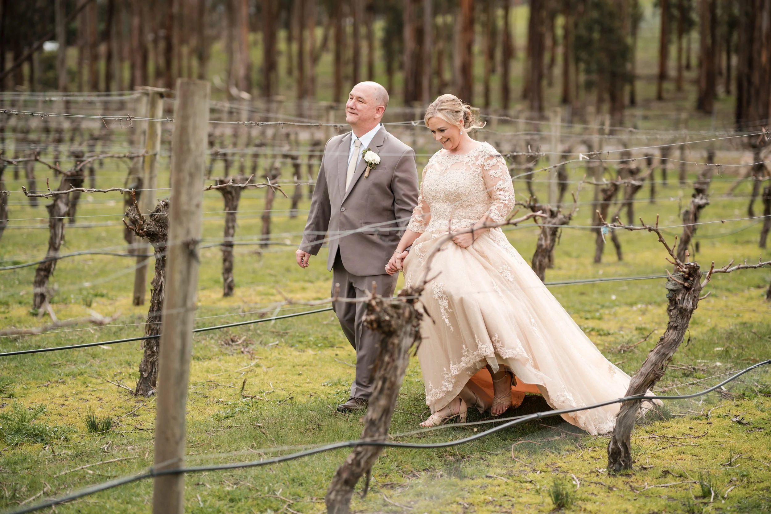 A bride and groom walking hand in hand through a vineyard.