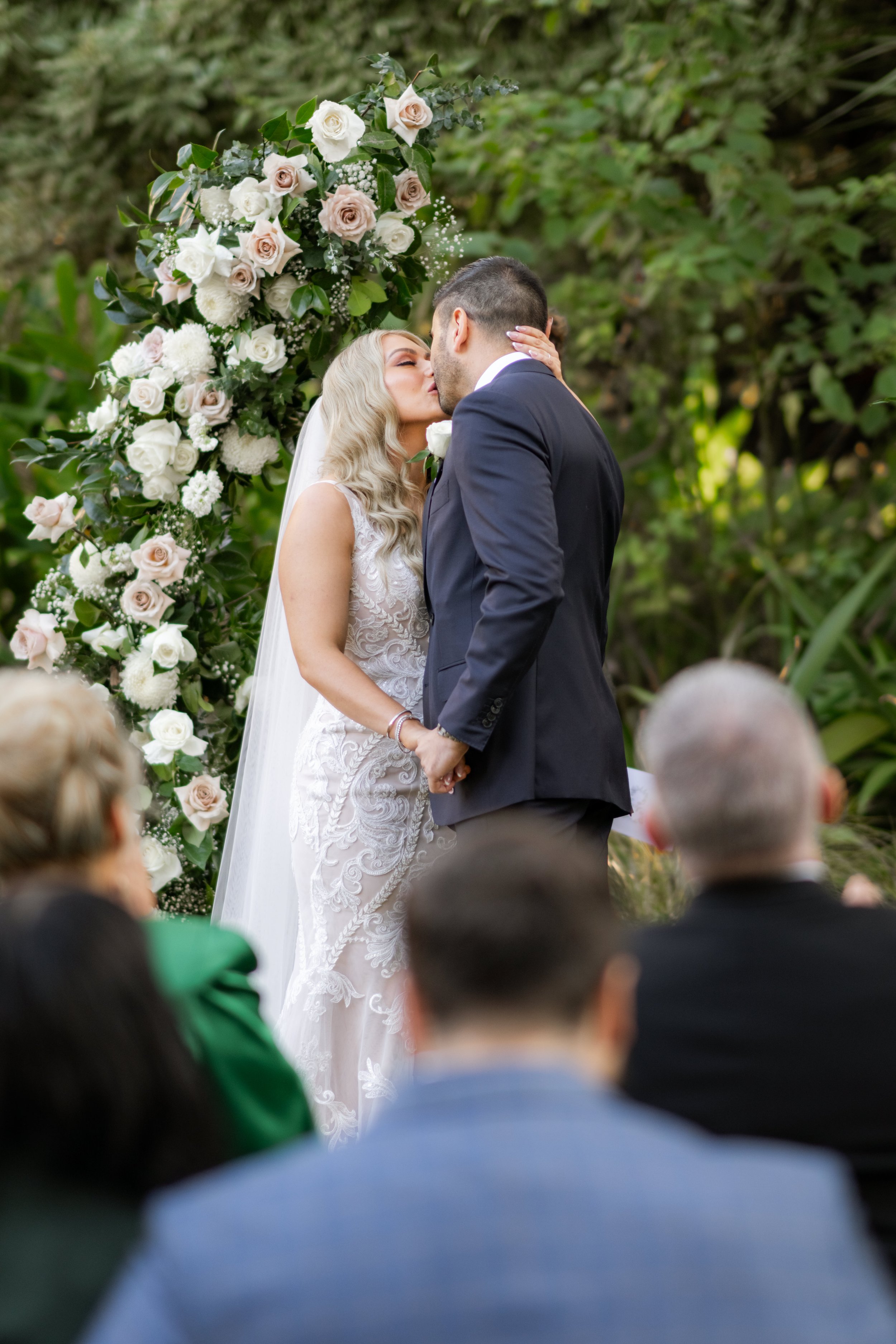 Bride and groom kissing at outdoor wedding ceremony, surrounded by floral arch and greenery.