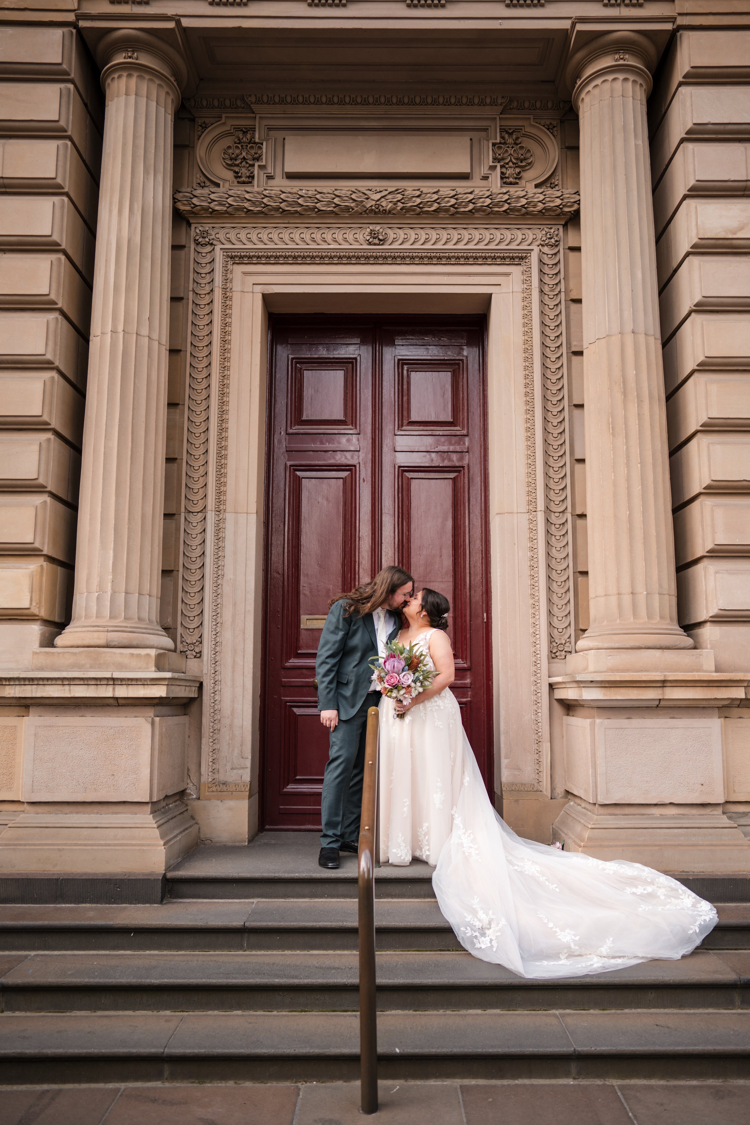 A couple dressed in wedding attire standing on steps in front of a large wooden door with ornate stone framing, sharing a kiss.
