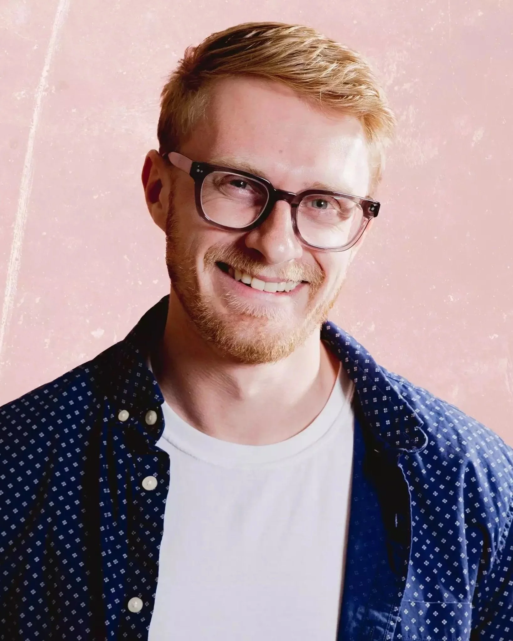 Close-up of a smiling man with red hair, beard, and glasses wearing a white T-shirt and a blue shirt with white polka dots, against a pink background.