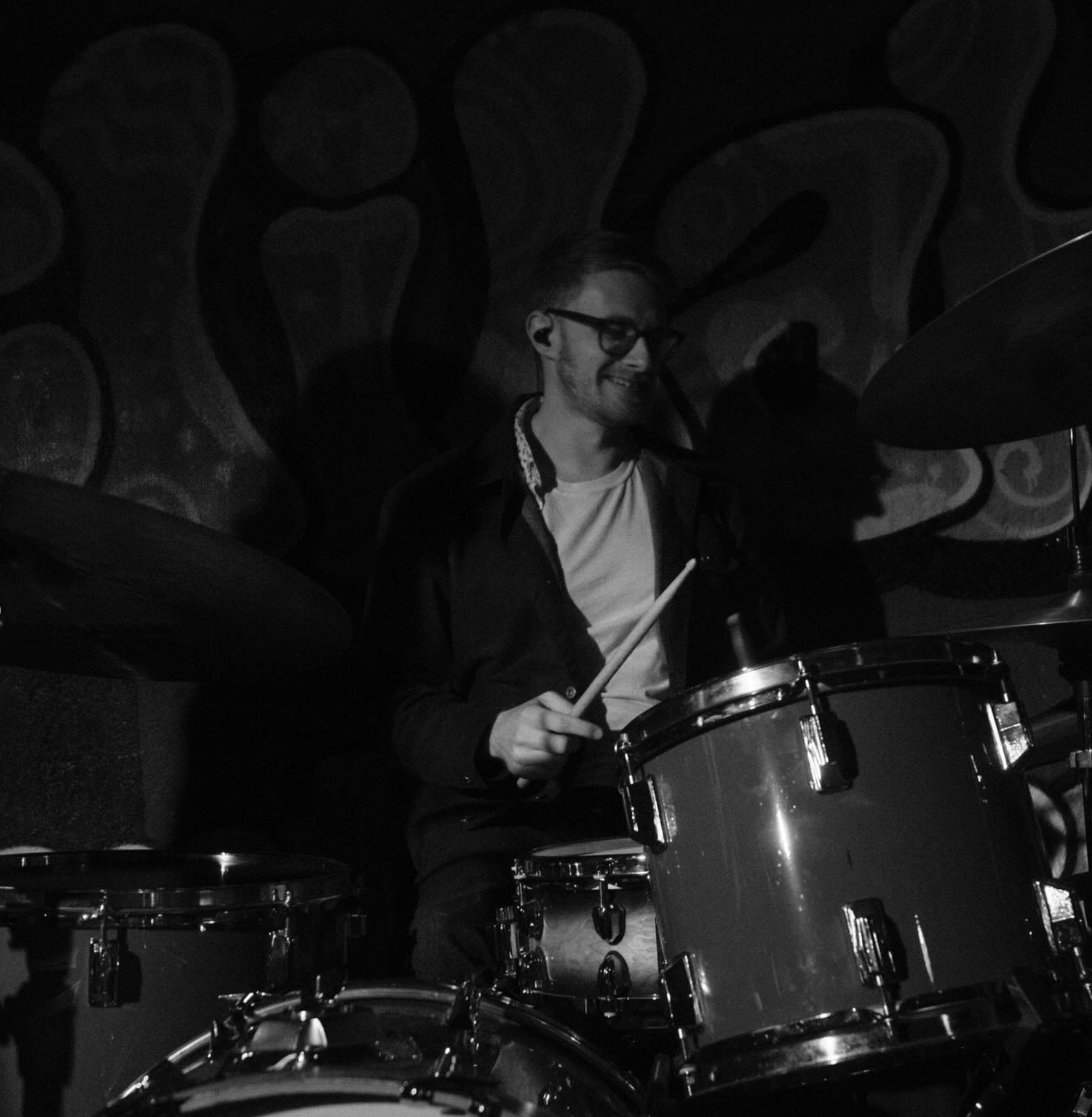 A young man with glasses playing drums, smiling, with a colorful graffiti wall in the background.