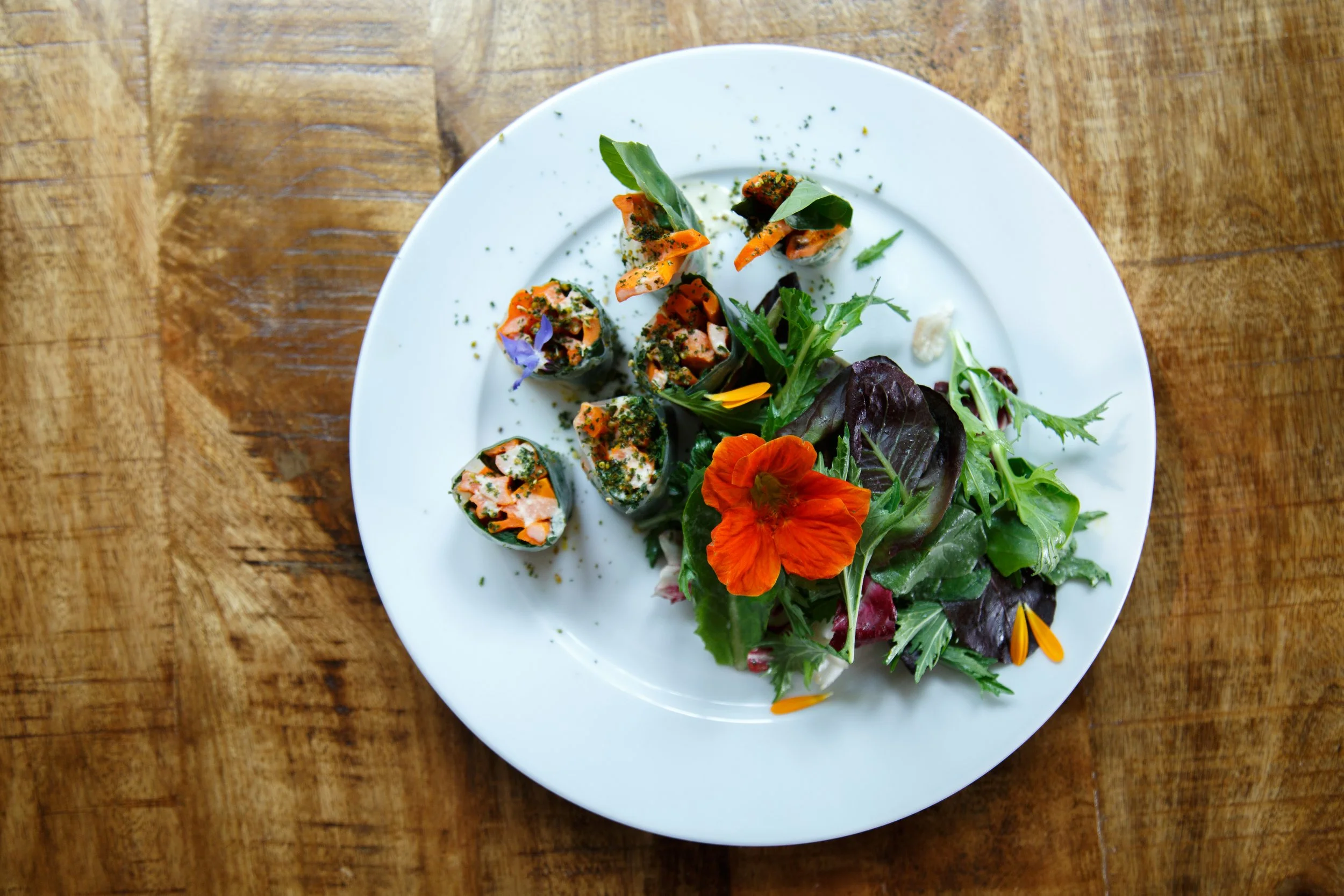 A white plate with a salad of mixed greens, edible flowers, and sliced vegetable rolls on a wooden table.