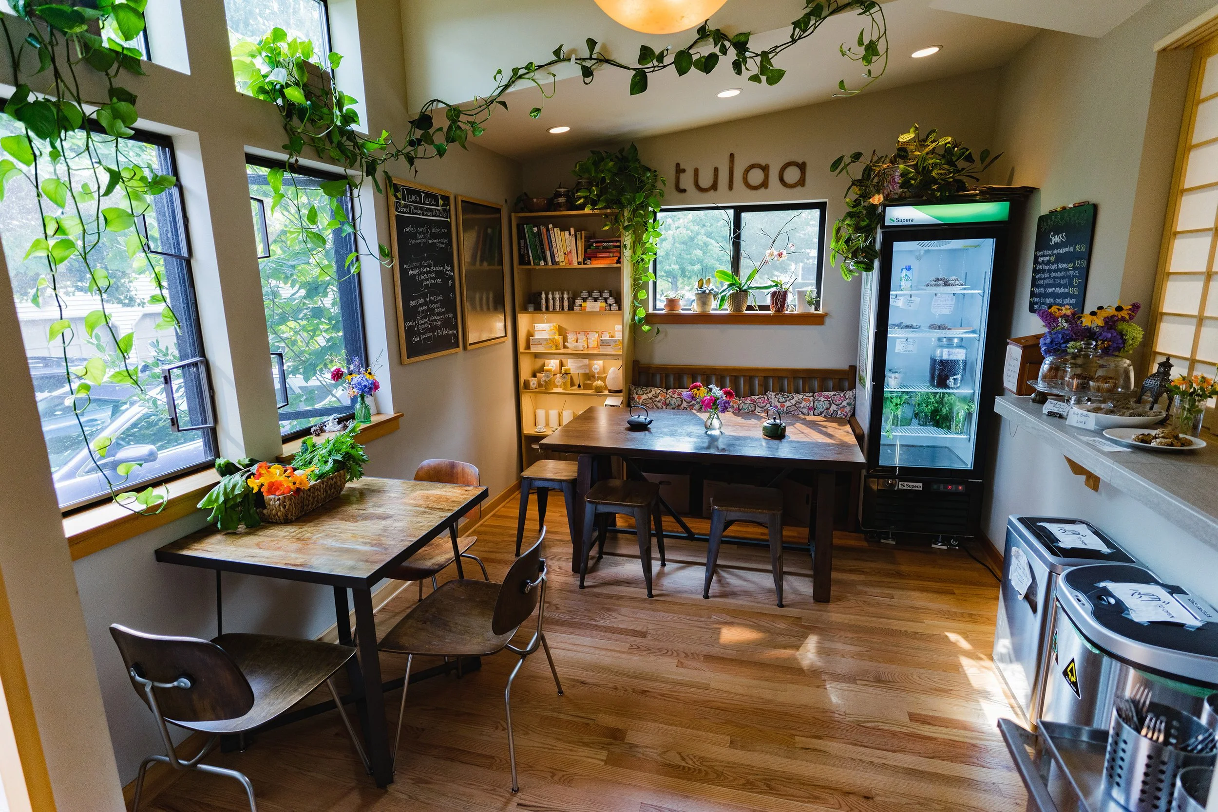 Cozy cafe interior with wooden tables and chairs, green plants on windowsills, a chalkboard menu, colorful flowers, a refrigerator with drinks, and a decorated counter area.