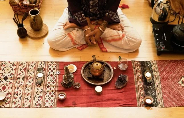 Person sitting cross-legged on the floor with a spiritual or meditation setup, including candles, a teapot, and crystals on a decorative cloth.