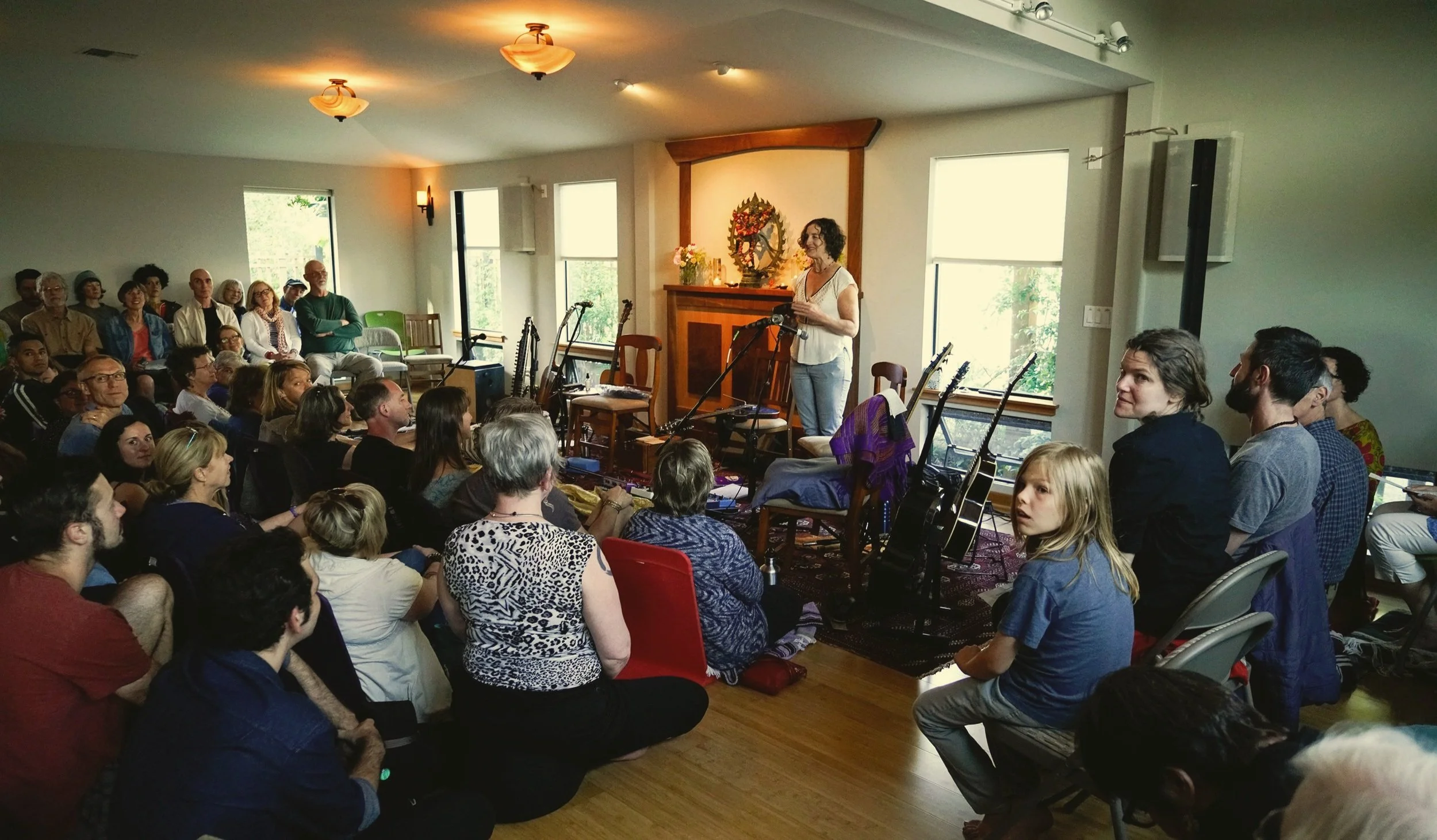 A woman is speaking on a stage with multiple guitars behind her, in front of an audience seated closely in a cozy, well-lit room.