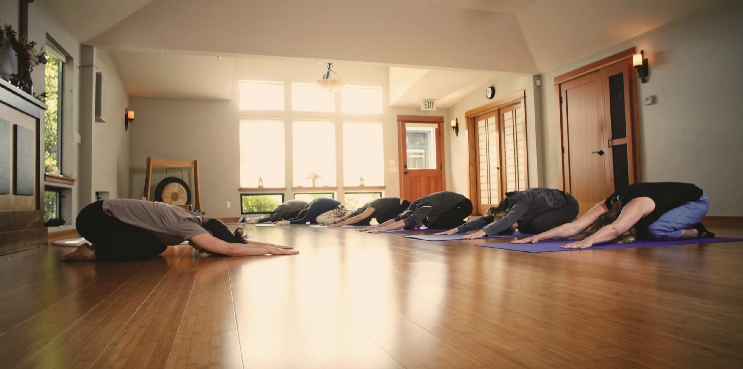 Group of people participating in a yoga class in a brightly lit room, practicing Child's Pose on purple mats.