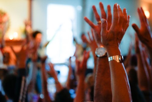 Multiple people raising their hands in a group indoor setting, during a yoga class.