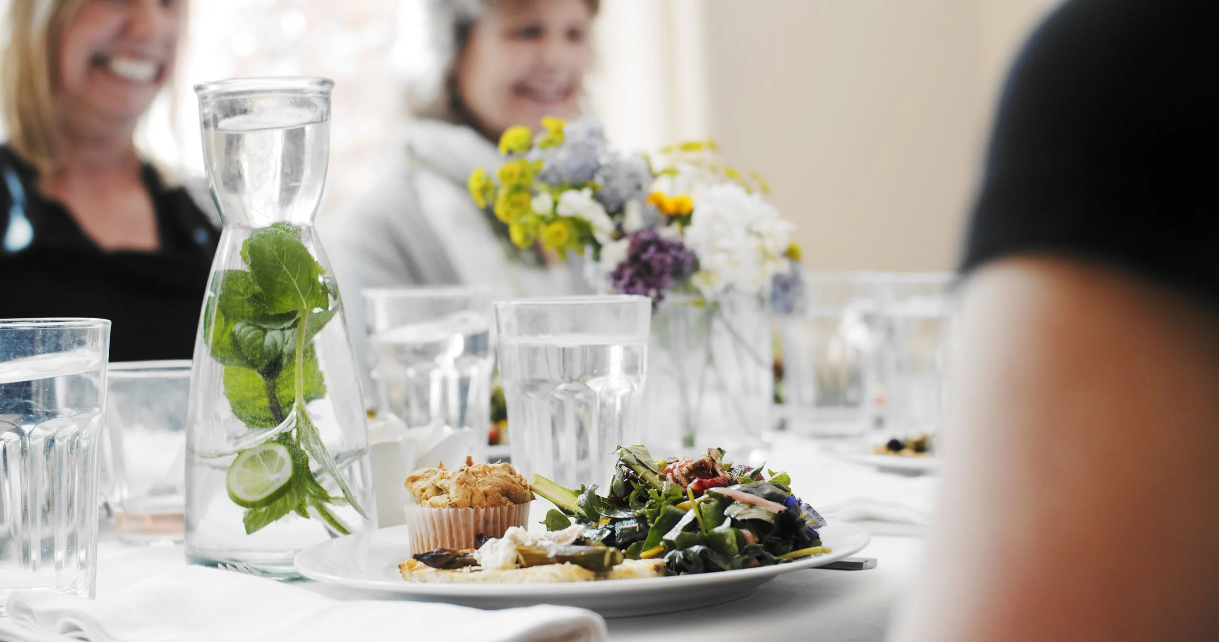 A dinner table with a glass water pitcher with mint and lime, plates with food, and a floral centerpiece, with smiling women in the background.