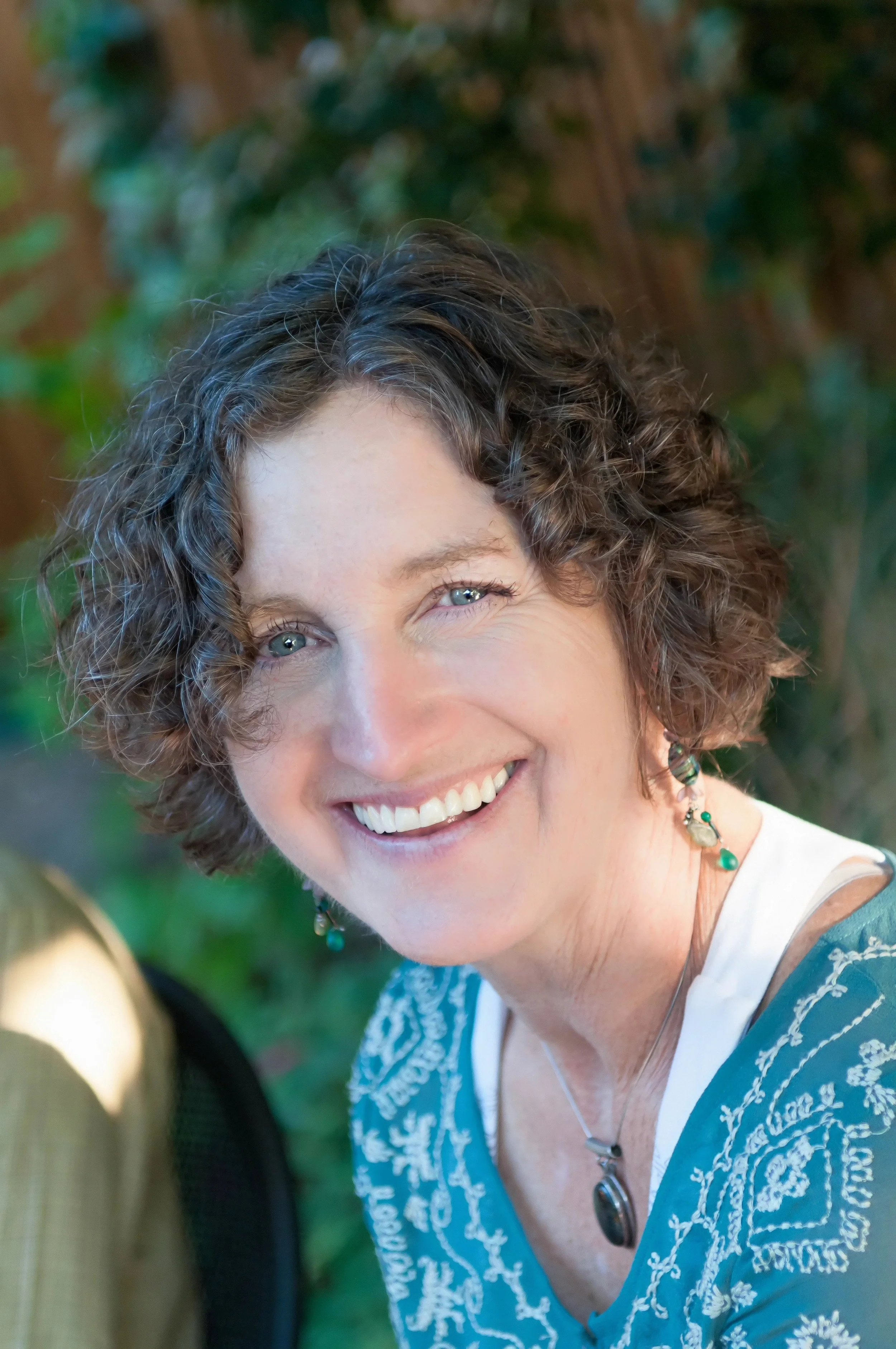 Close-up of a smiling woman with short curly hair, wearing a teal embroidered top and jewelry, outdoors with a blurred green background.