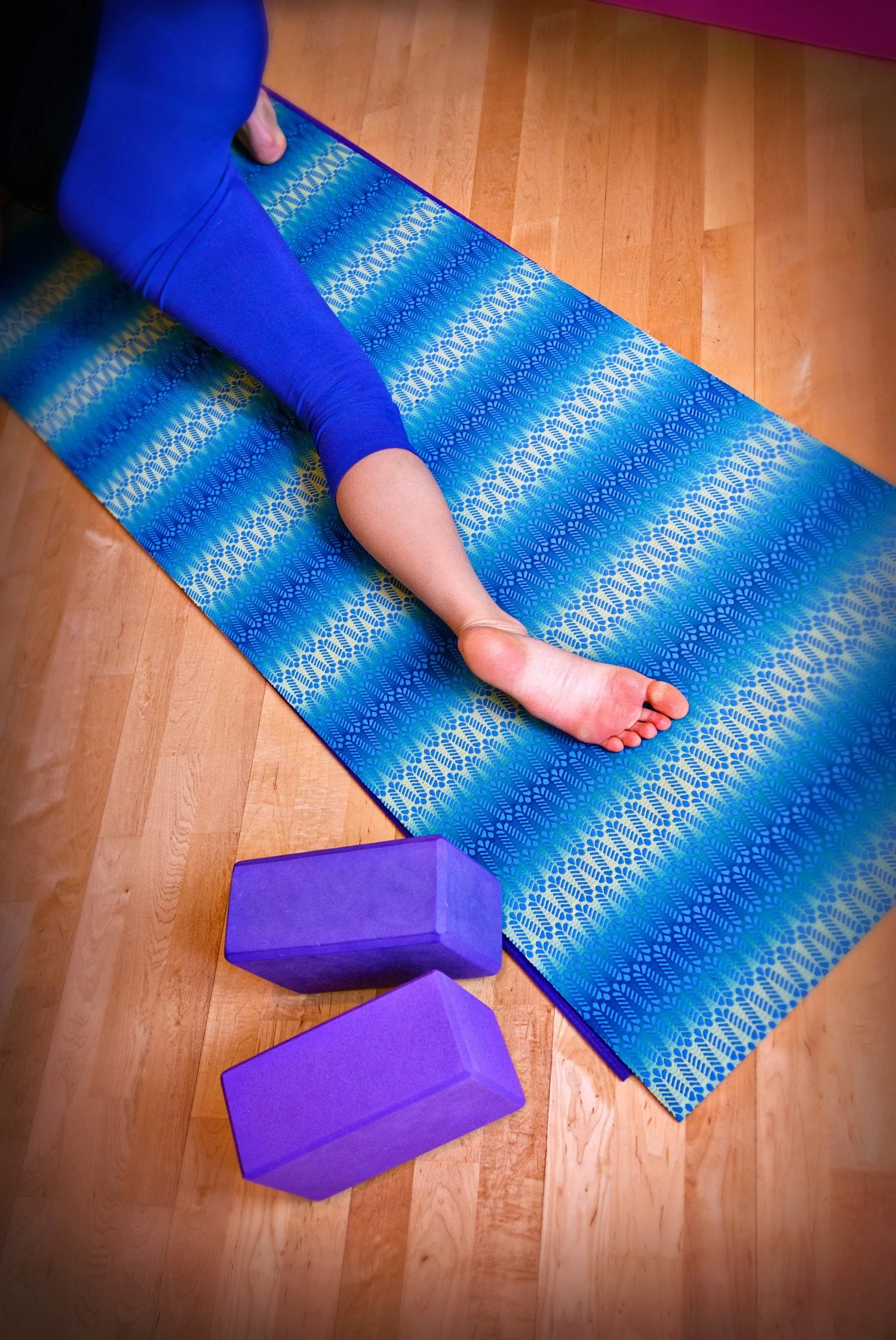 Person doing stretching or yoga on a blue patterned mat wearing blue leggings, with two purple yoga blocks nearby on a wooden floor.