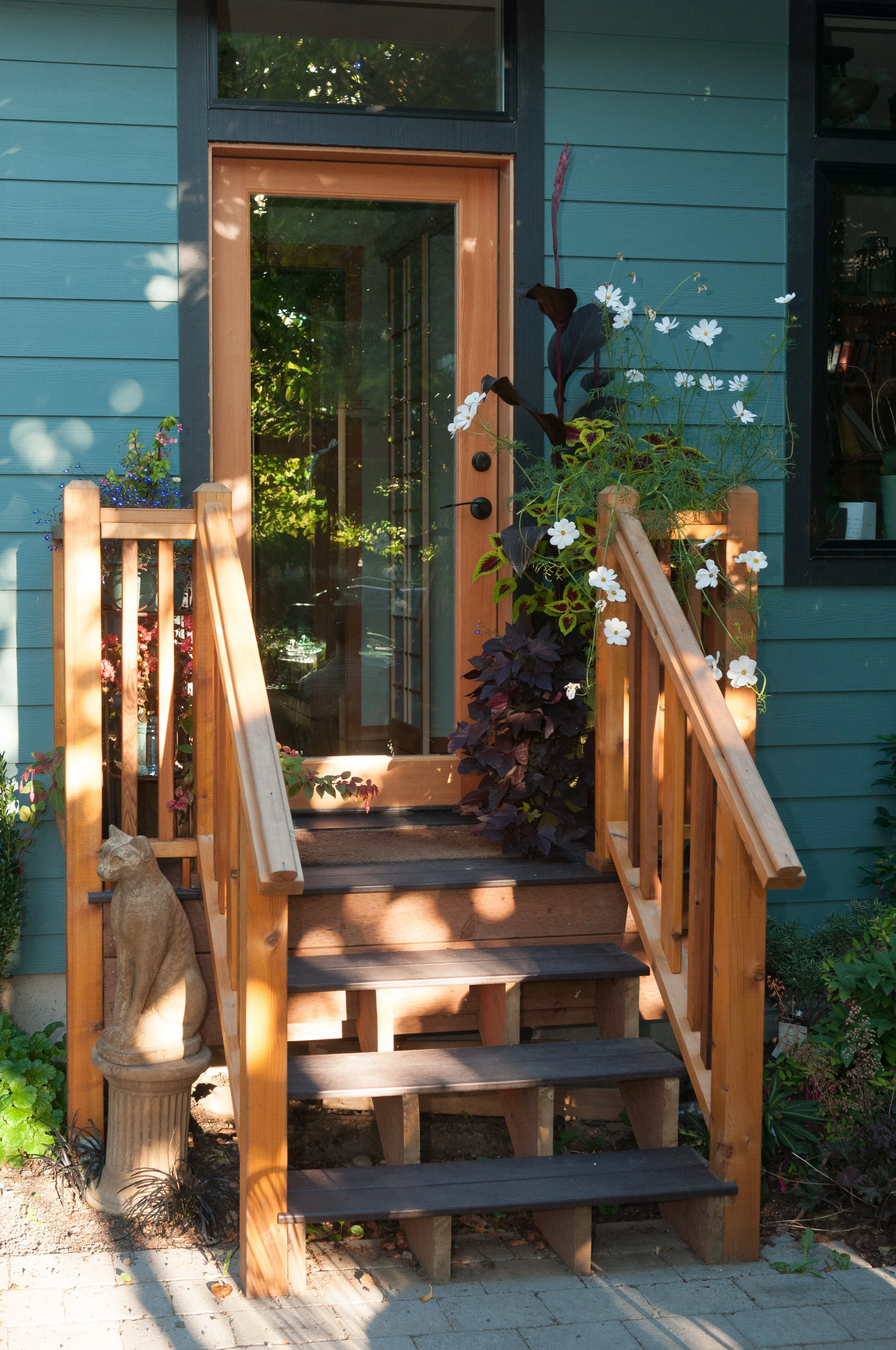 Wooden front steps leading to a glass door on the teal-colored studio, decorated with potted plants and a cat statue on a pedestal by the stairs.