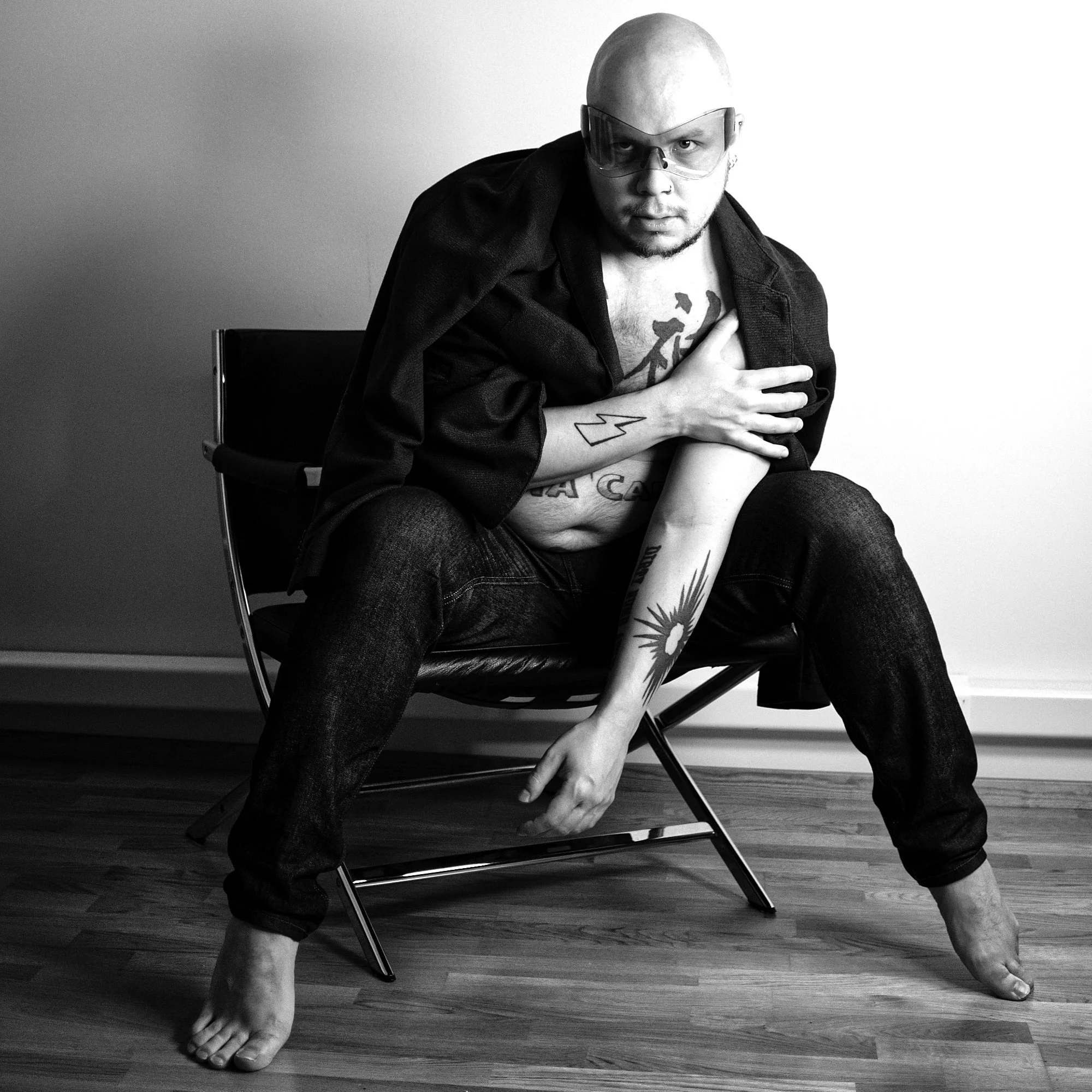 Black and white photo of Peter Juhan, wearing glasses, sitting on a chair with a wide stance. He is shirtless with tattoos visible on his arm and chest, and he is wearing a open jacket and jeans.