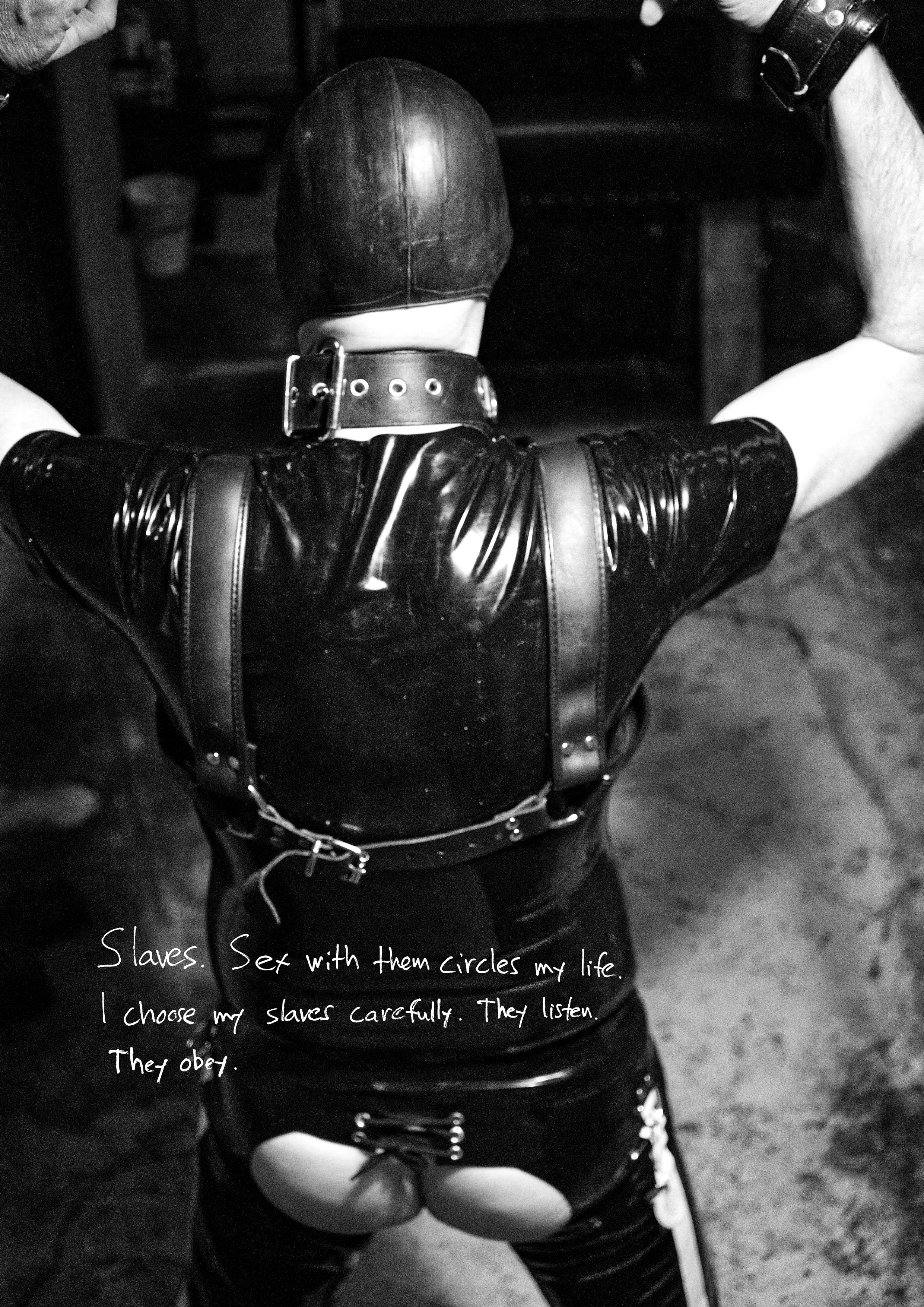 Person dressed in leather, wearing a mask and harness, with hands raised and held by two individuals. There is handwritten text on the photo that says, "Slaves. Set with them circles my life. I choose my slaves carefully. They listen. They obey."