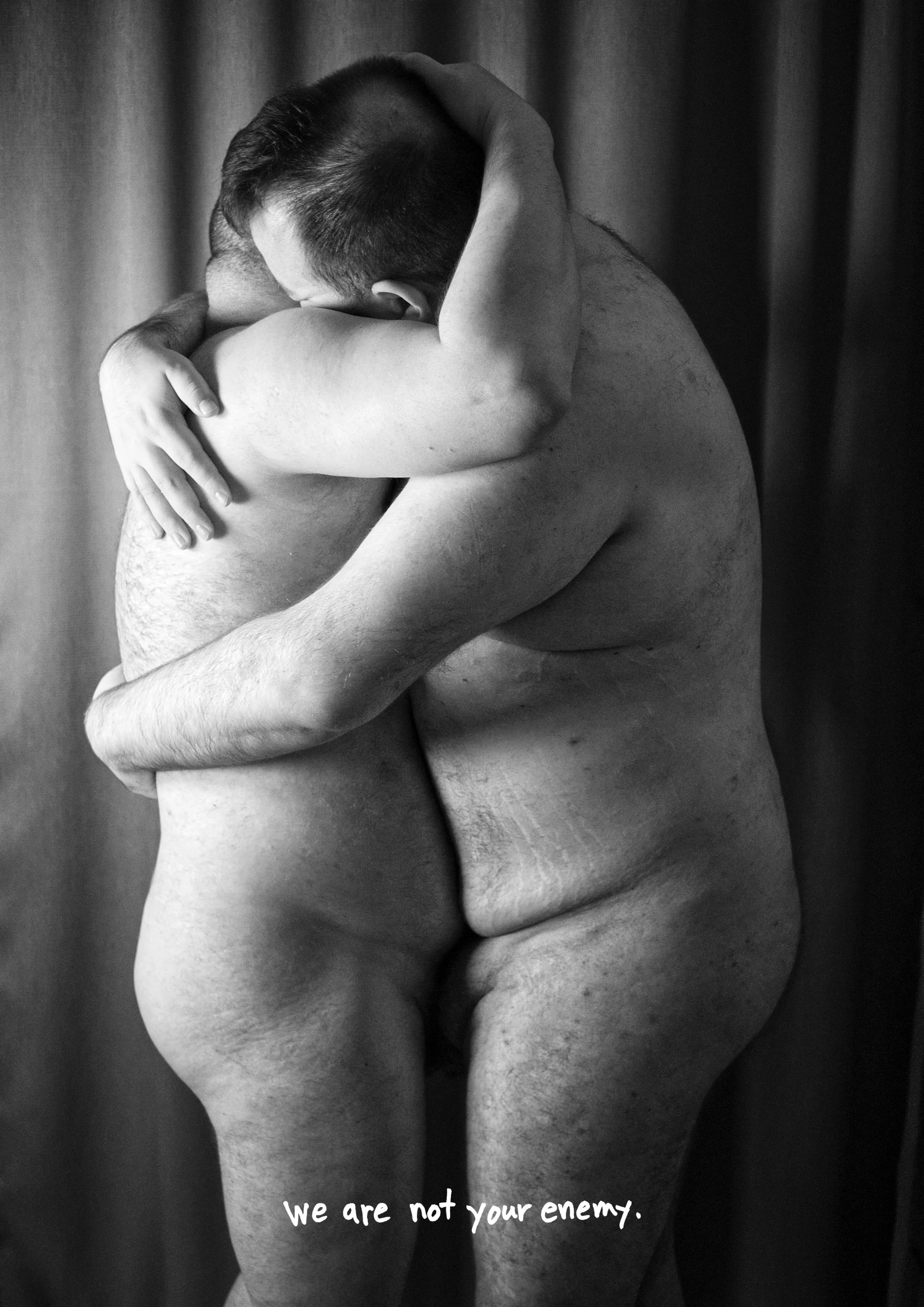 Two naked men hugging each other in an intimate embrace with their faces pressed together. The background features a curtain, and there is text at the bottom that reads, "We are not your enemy."