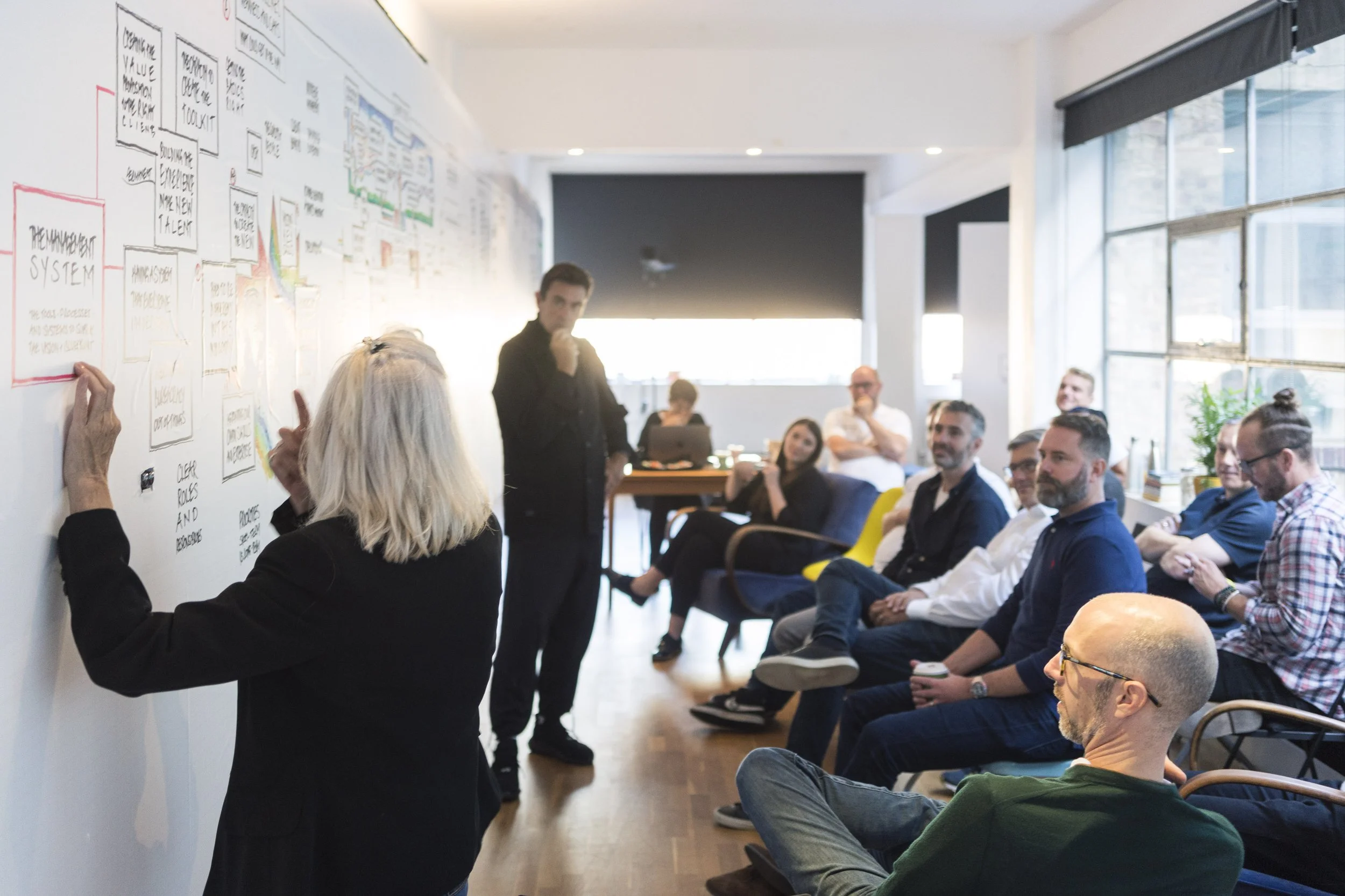 A group of people attending a presentation in a bright room with large windows. A woman is standing at a whiteboard filled with notes and diagrams, addressing the seated audience.
