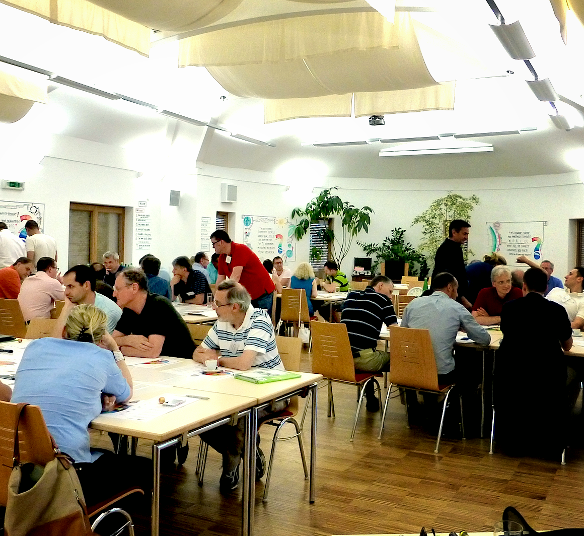 People seated at tables in a conference or workshop room with white walls, wooden floors, and large plants in the background.