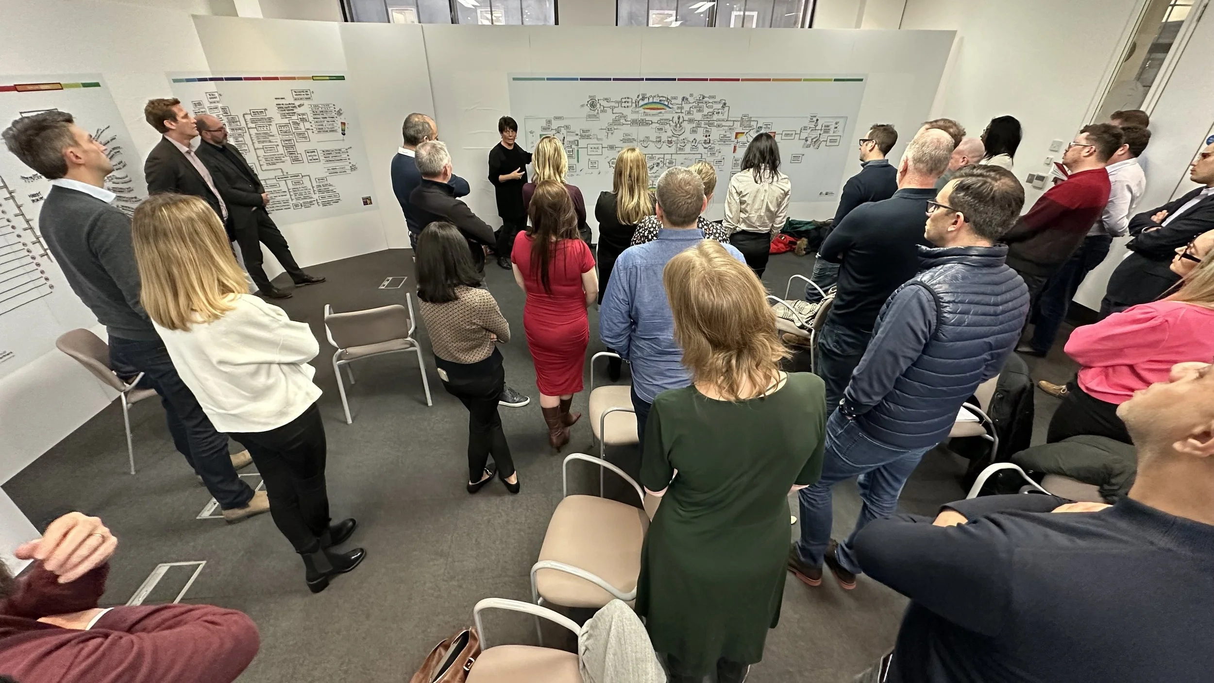 A woman presenting to a group of people in a conference room. The audience is standing and seated, listening attentively. The room has white walls with charts and diagrams.