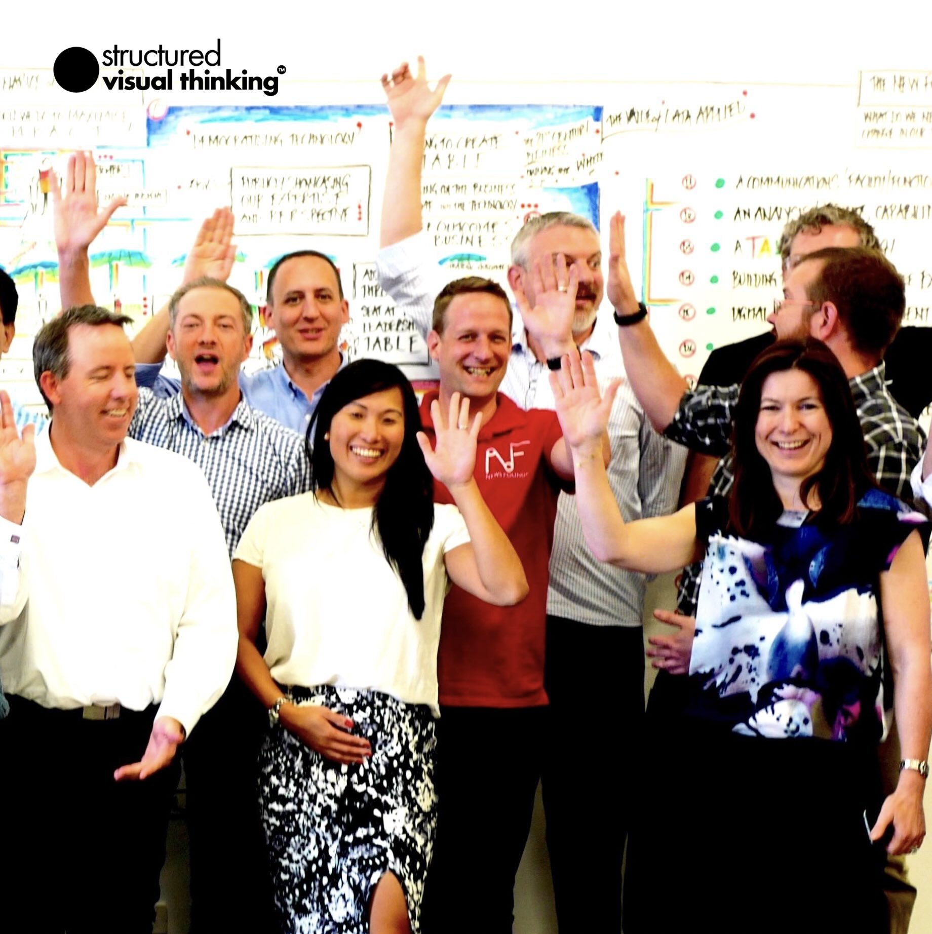 Group of people smiling and waving next to a whiteboard with text and drawings. The image includes men and women, and one person is wearing a 'structured visual thinking' shirt.