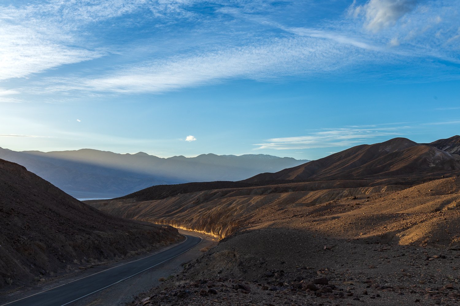 A bend in the road on Artists' Drive, Death Valley, California. Blue sky with clouds above, and the mountains glowing with golden hour light.