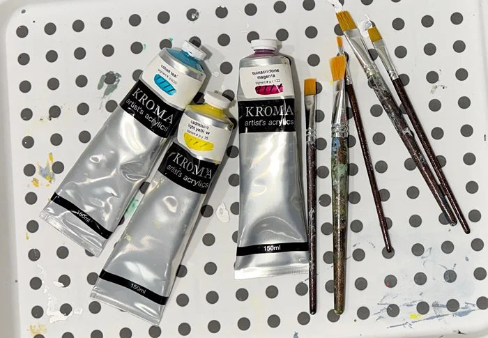 Tubes of teal, yellow and magenta paint on a tray with small brushes.