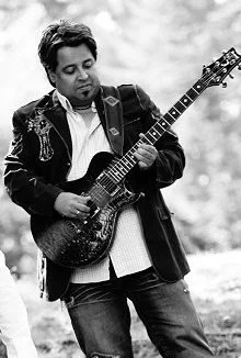 The Owner of Boca Music, playing an electric guitar outdoors, focused on strumming the instrument.