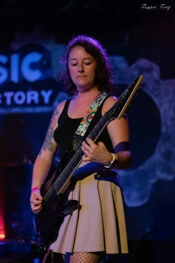 Young woman playing an electric bass guitar on stage, wearing a black sleeveless top, beige skirt, and fishnet stockings, with a dark background featuring blue text.