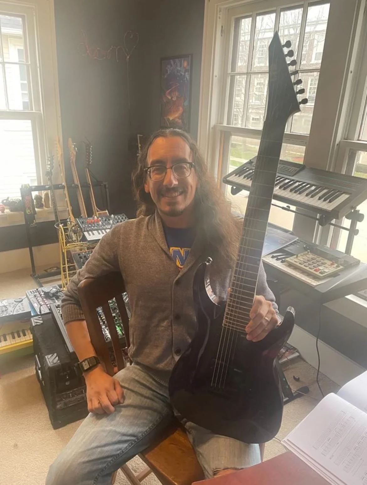A man with long hair, glasses, and a beard smiling while holding a black electric guitar in a music studio filled with musical instruments and equipment.