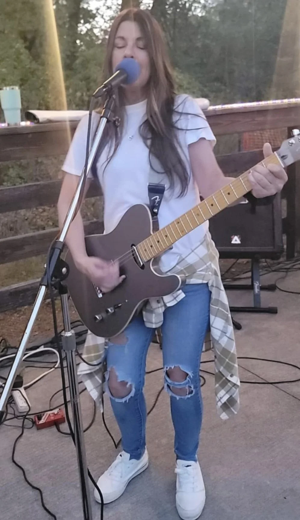 A young woman with long brown hair is singing into a microphone while playing an acoustic guitar outdoors. She wears a white t-shirt, ripped jeans, white sneakers, and has a plaid shirt tied around her waist.