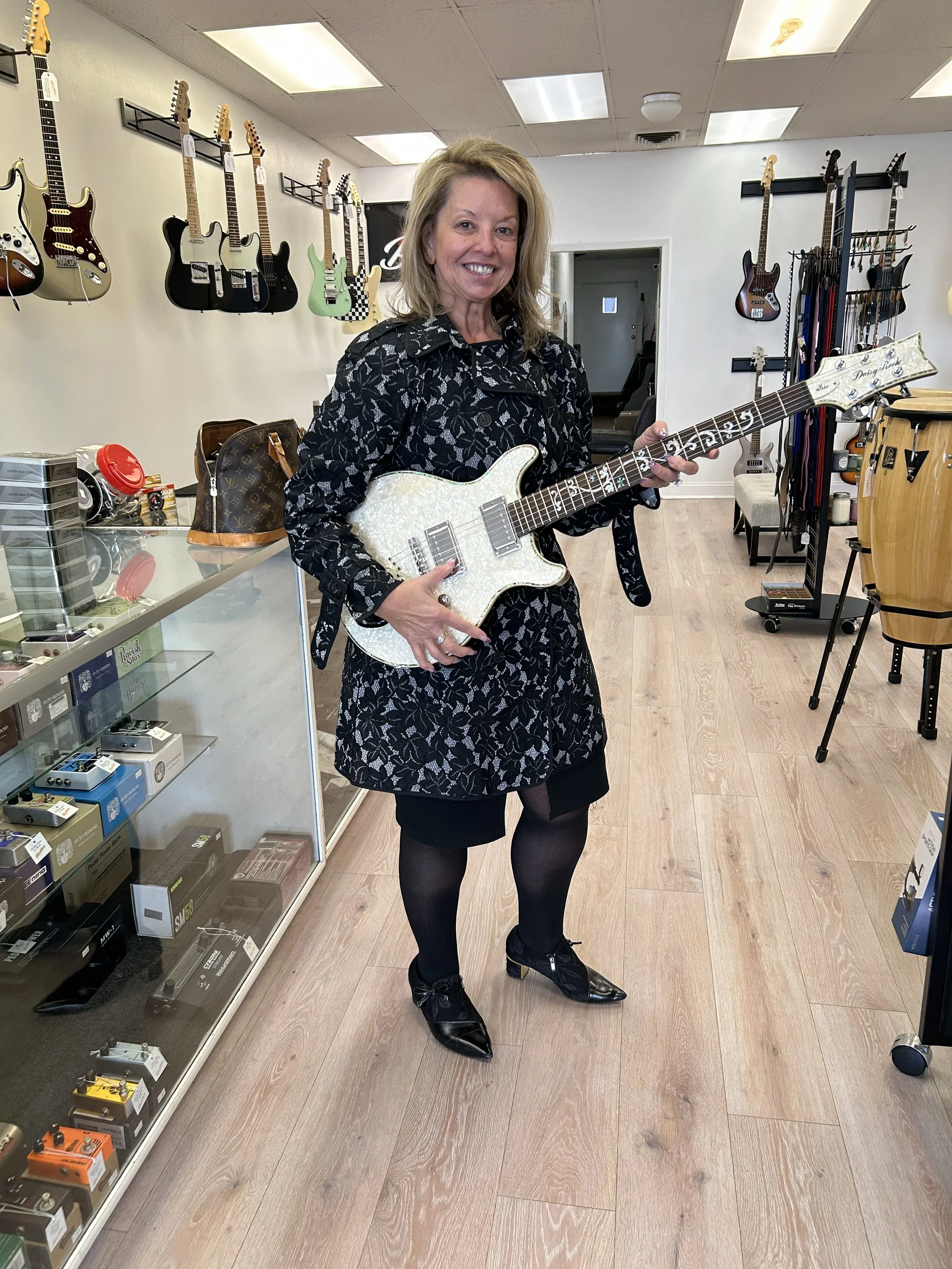 A woman standing inside a music store holding a white guitar with a pearl-like finish. The store has various guitars hanging on the wall behind her and music equipment on display.