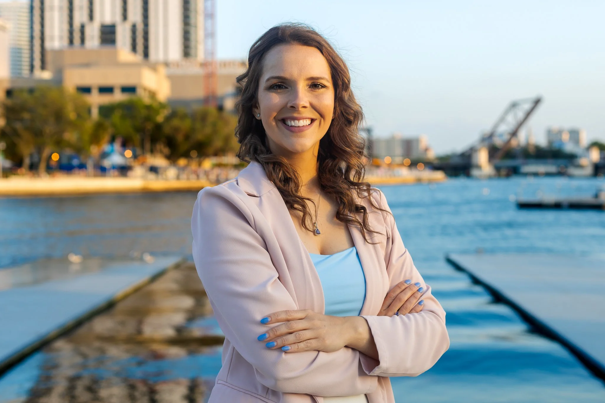 A woman with brown curly hair and fair skin standing by water with a city skyline in the background, smiling with arms crossed, wearing a light pink blazer and a blue top.