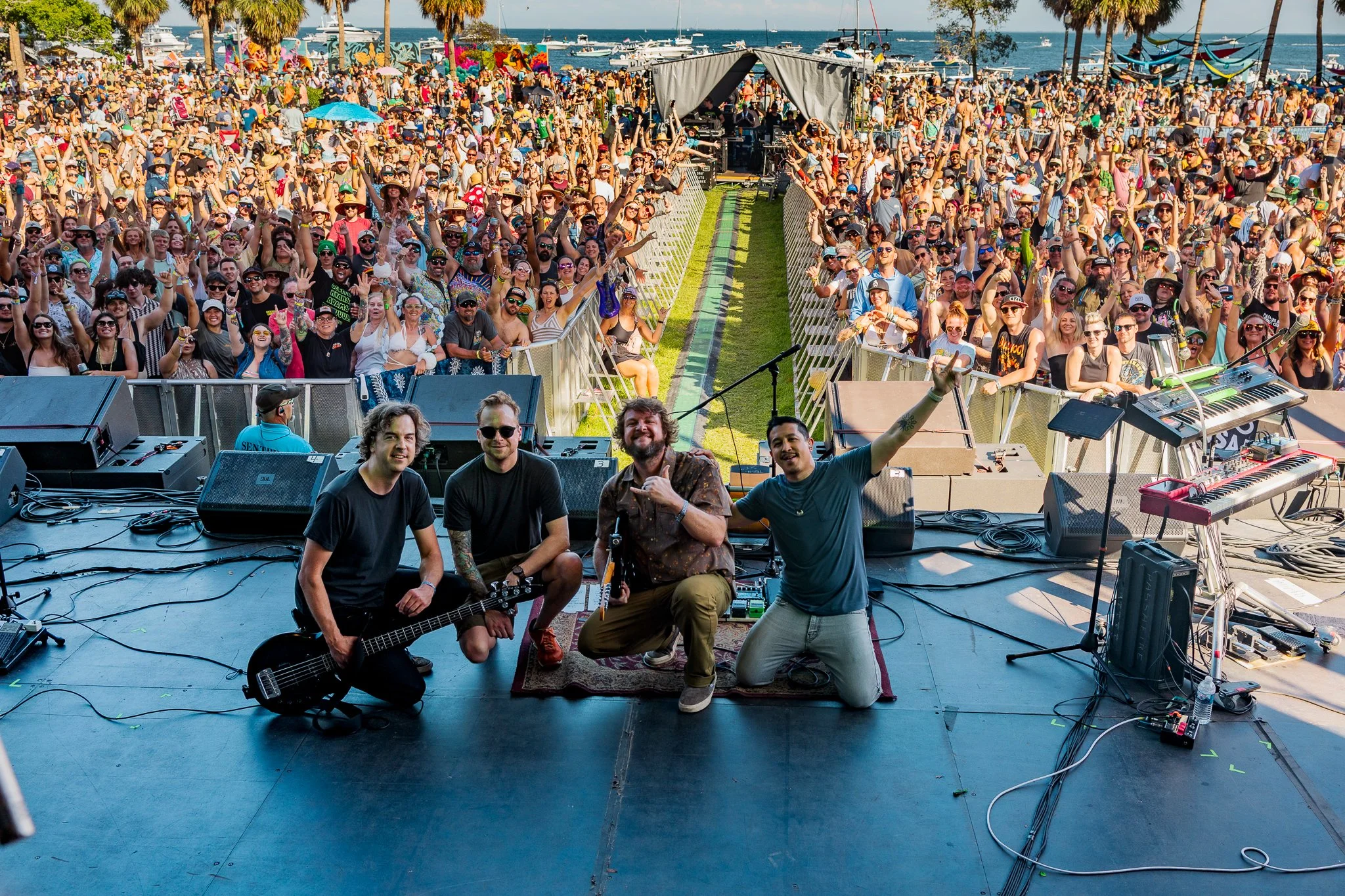 A band performing on stage at an outdoor concert by the water, with a large crowd of festival attendees enjoying the music under sunny skies.