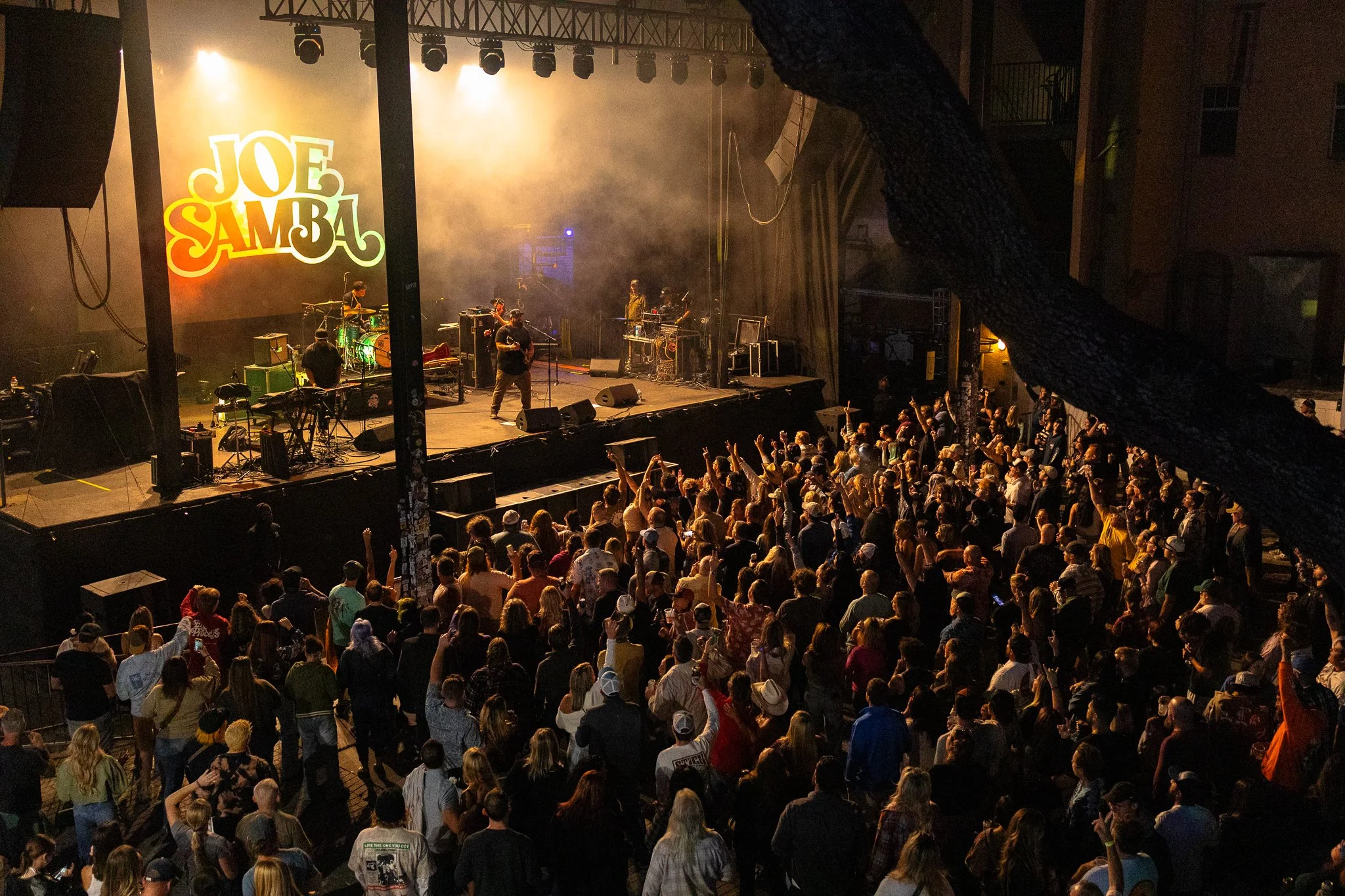 A crowd of people gathered in front of a stage at a concert, with band performing and a large screen displaying the name "Joe Samba" in colorful lettering.