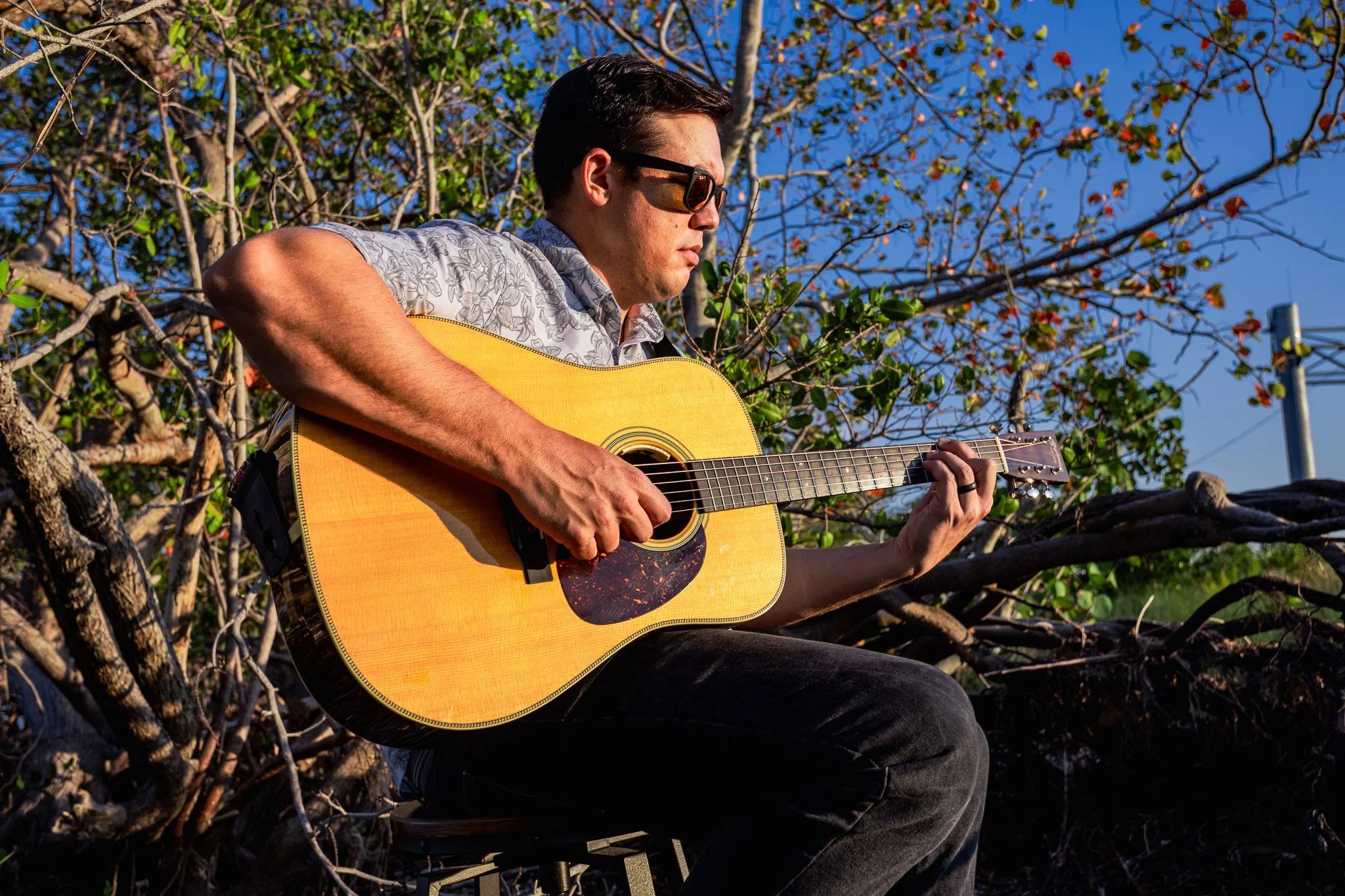 A man wearing sunglasses playing an acoustic guitar outdoors among tree branches on a sunny day.