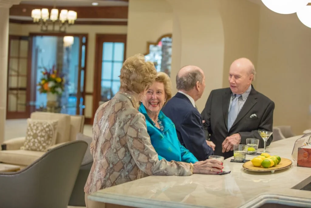 Four older adults smiling and talking together around a counter in a shared indoor space.