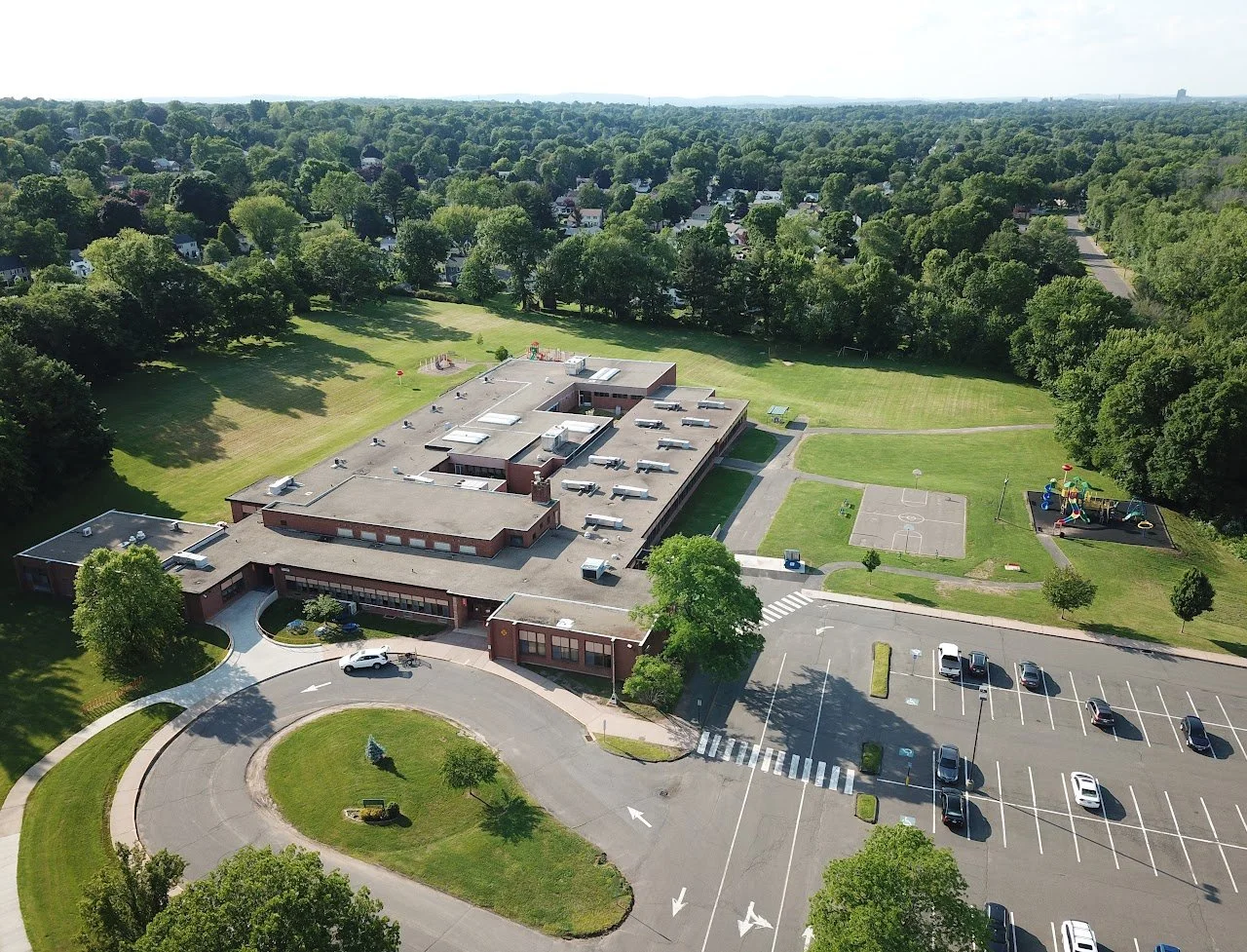 Aerial view of a school building with a parking lot, playground, basketball court, and extensive green grassy areas surrounded by trees.