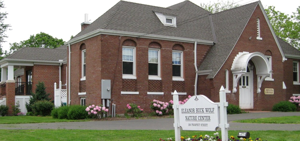 A red brick building with a white sign in front that reads Eleanor Buck Wolf Nature Center, located at 150 Prospect Street, with a well-maintained green lawn and pink flowers