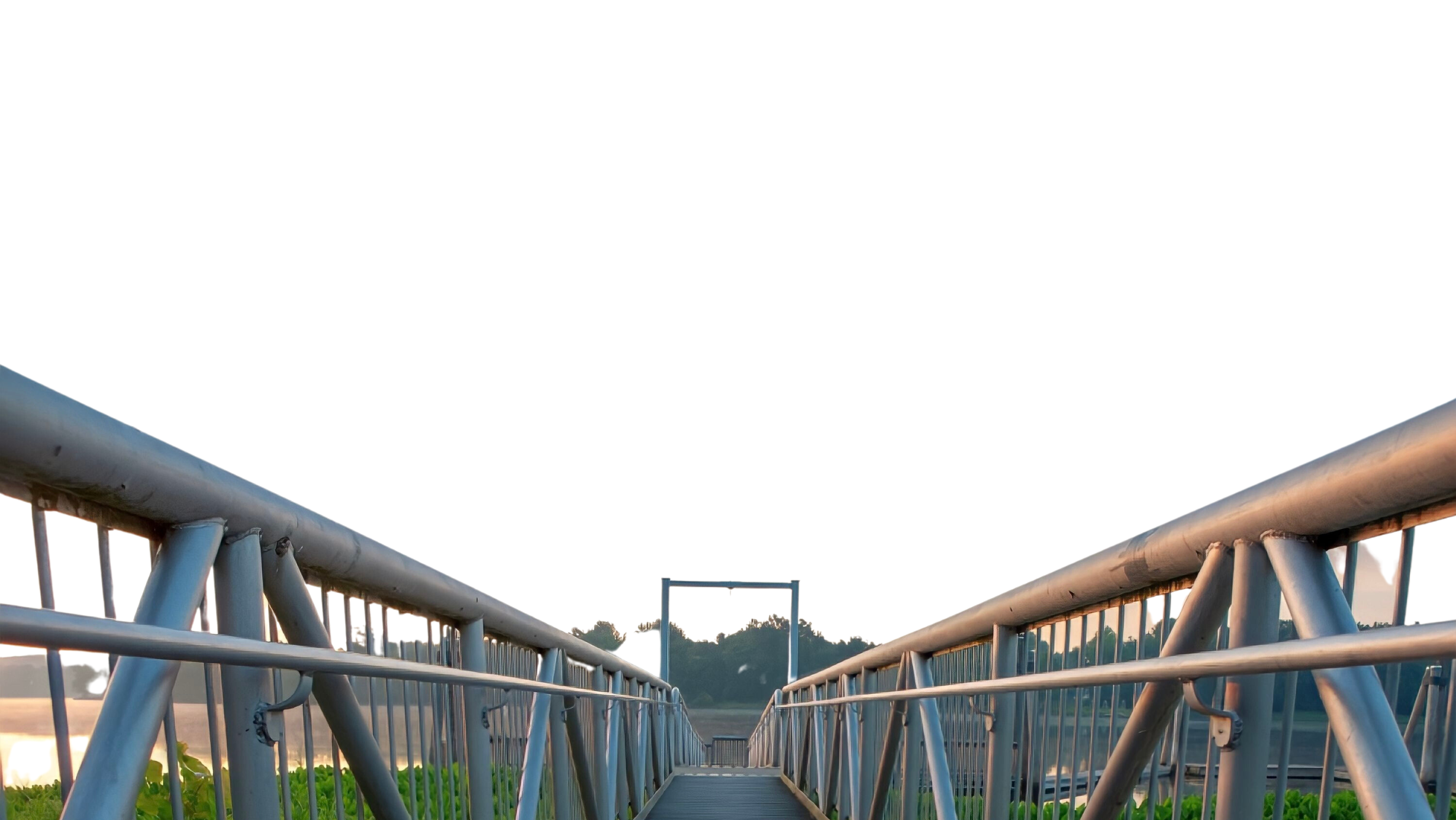 View of a metal bridge or walkway with railings, leading towards a distant gate or frame, person or object is not visible, with trees and a body of water in the background at sunset or sunrise.