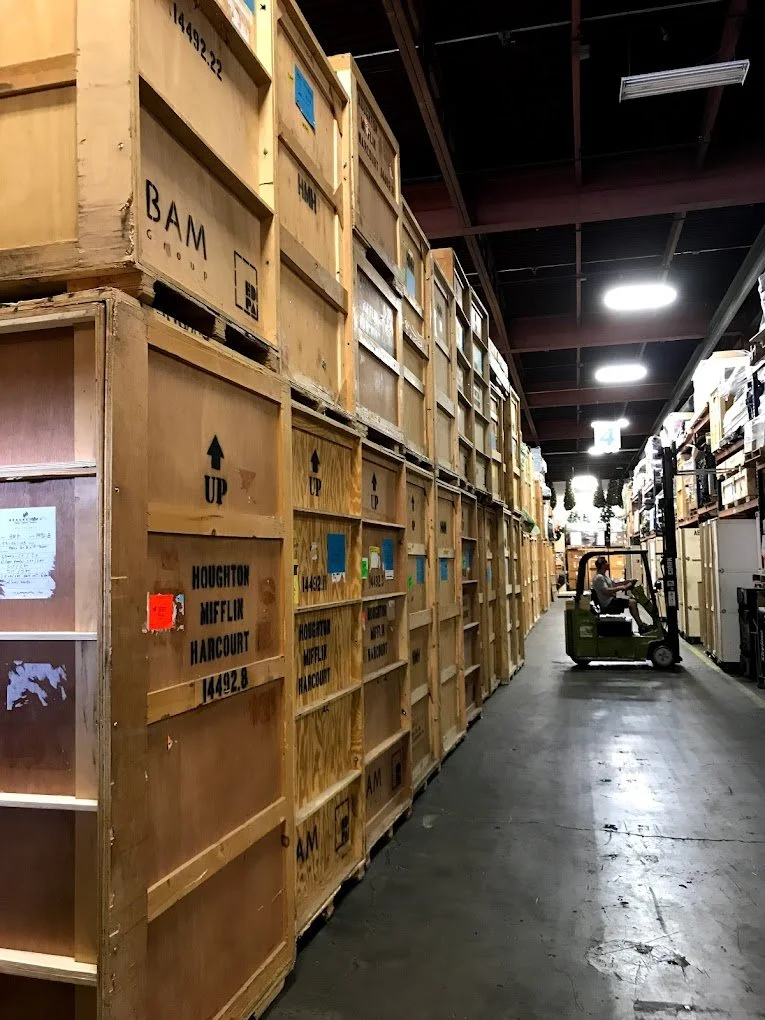 Warehouse aisle with tall stacks of wooden crates on the left and a forklift operator moving pallets on the right, high ceiling with overhead lights.