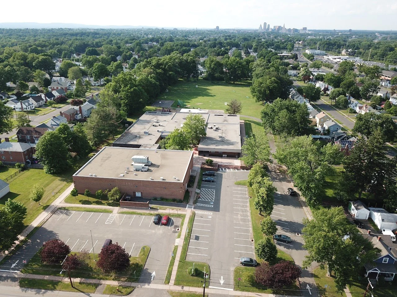 Aerial view of a school building with a large parking lot, surrounded by a residential neighborhood with houses and trees, and a green park area in the background.