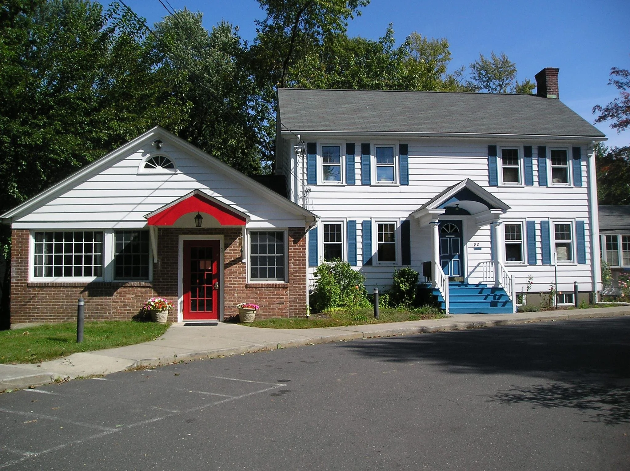 A white multi-story house with blue shutters, a small porch, and a gabled section with a red door at the front, surrounded by green trees and a paved parking area.