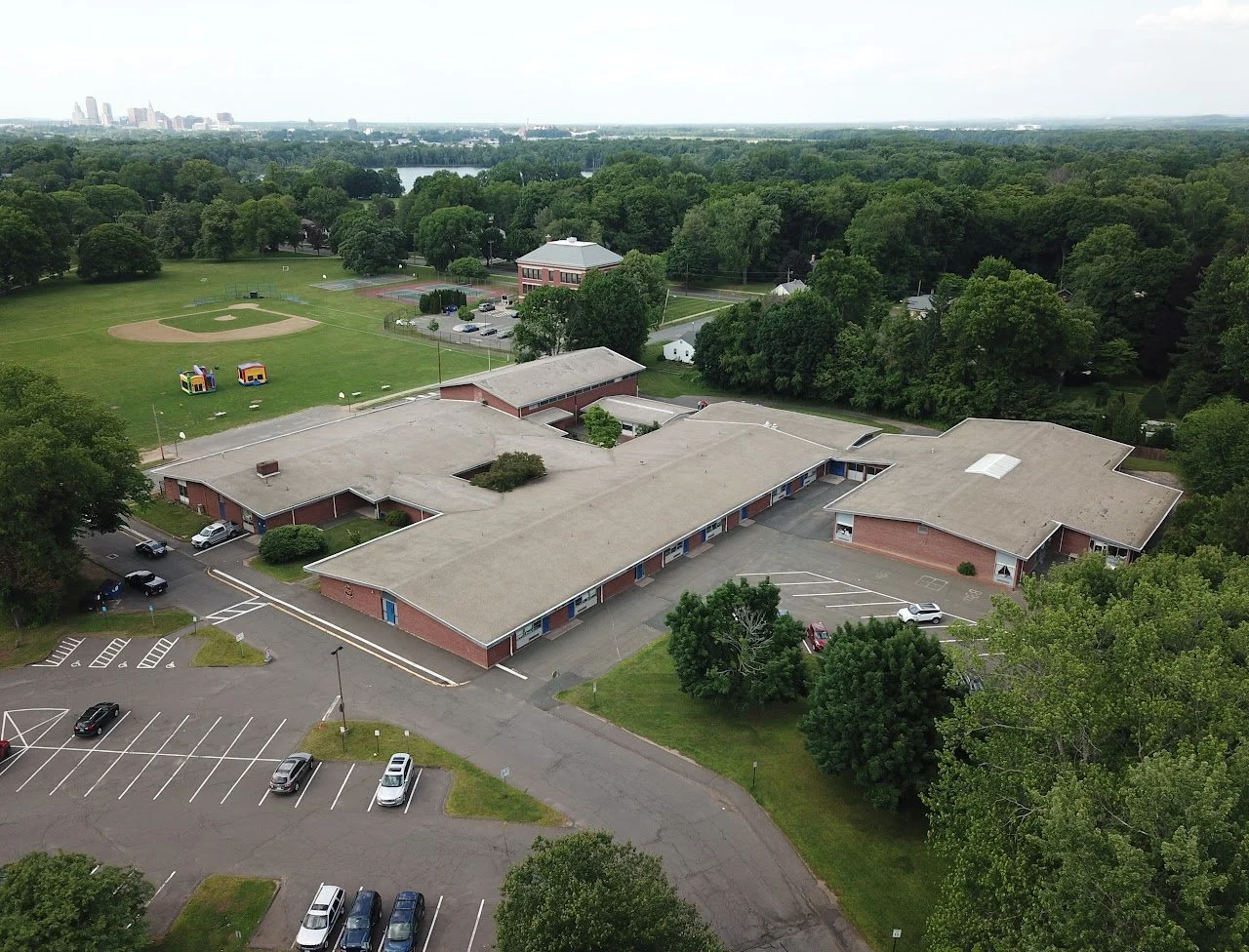 Aerial view of a school complex with parking lots, trees, a baseball field, and a community playground, with forested areas and a city skyline in the distance.